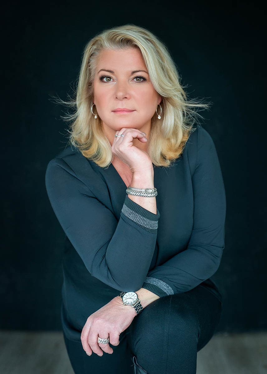 Blonde woman in dark clothing posing against a dark background with her hand resting near her chin, wearing silver jewelry and a watch.