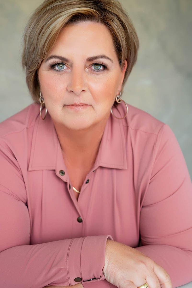 Woman with short light brown hair and blue eyes wearing a pink button-up shirt and hoop earrings, looking directly at the camera with a neutral expression and resting her arms on a surface.
