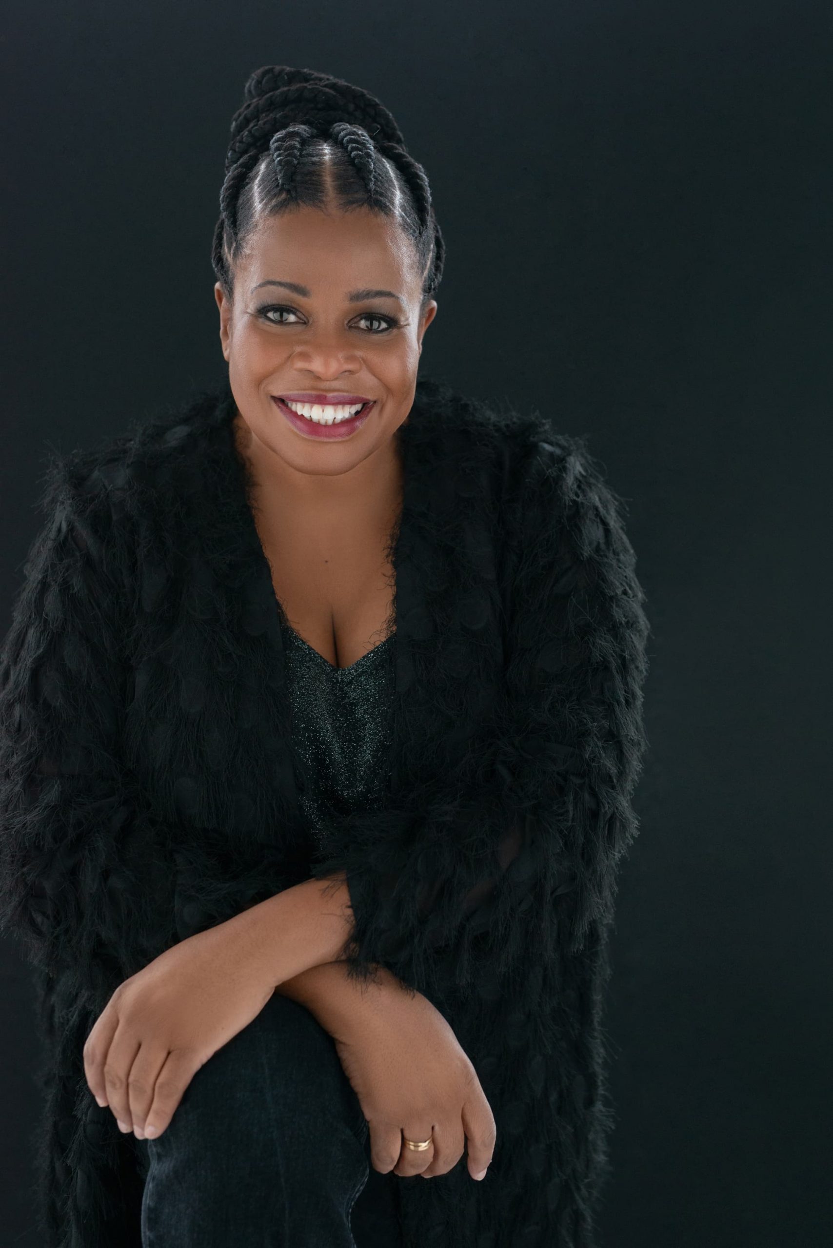Smiling Black woman with braided hair wearing a textured black coat and black top, seated with arms crossed over her knee against a dark background.