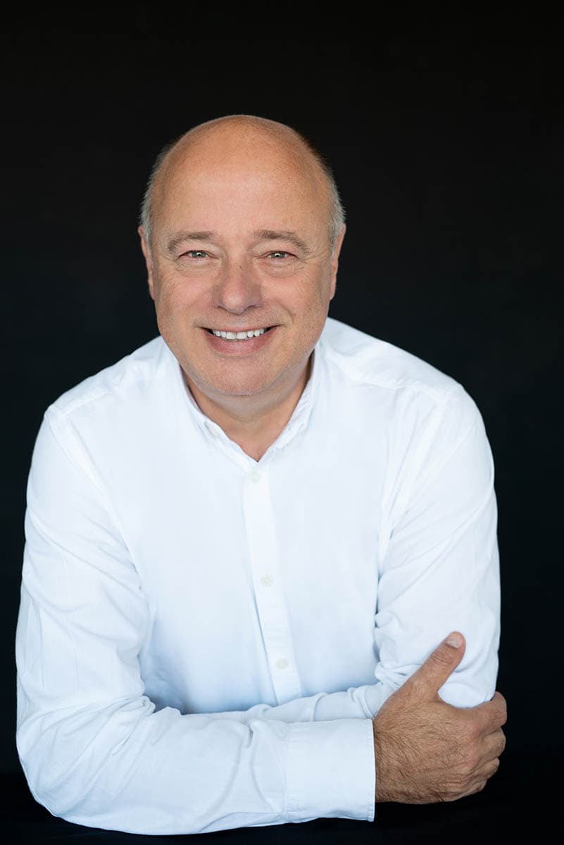 A middle-aged bald man smiling directly at the camera, wearing a white button-up shirt, with arms crossed and a black background.