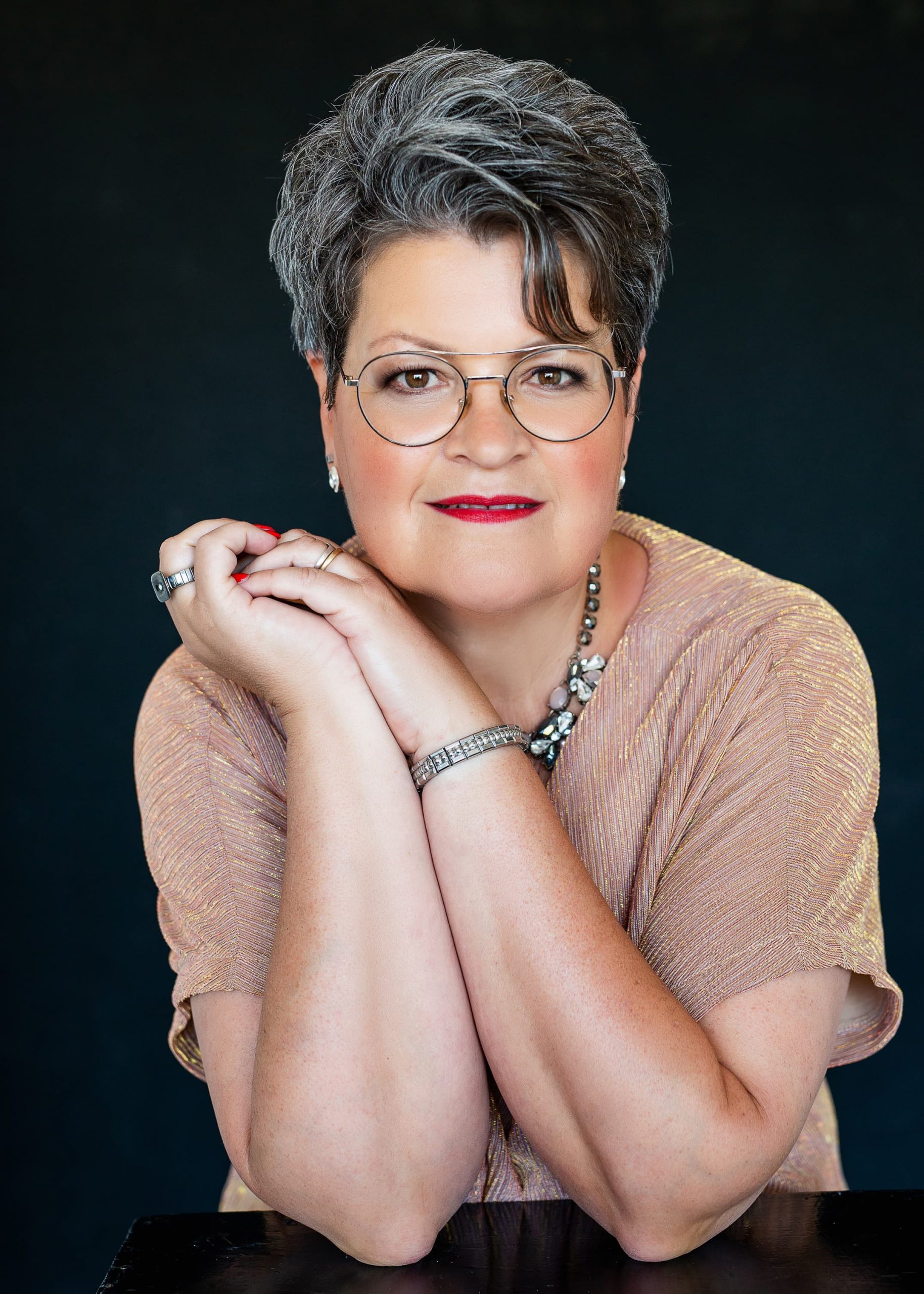 Middle-aged woman with short gray hair, wearing round glasses and a light textured top, resting her crossed arms on a black surface and looking at the camera with a slight smile.
