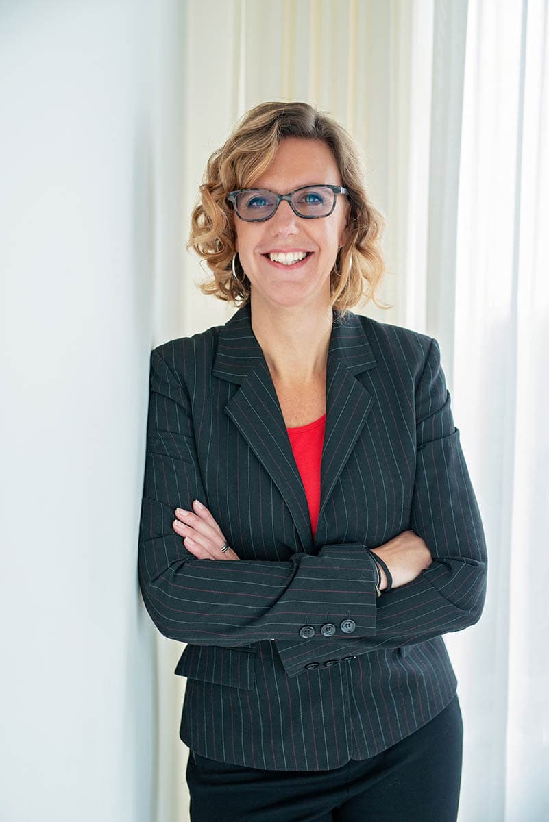 Smiling woman with curly blond hair wearing glasses, a black pinstripe blazer, and a red top stands with arms crossed in front of light-colored curtains.