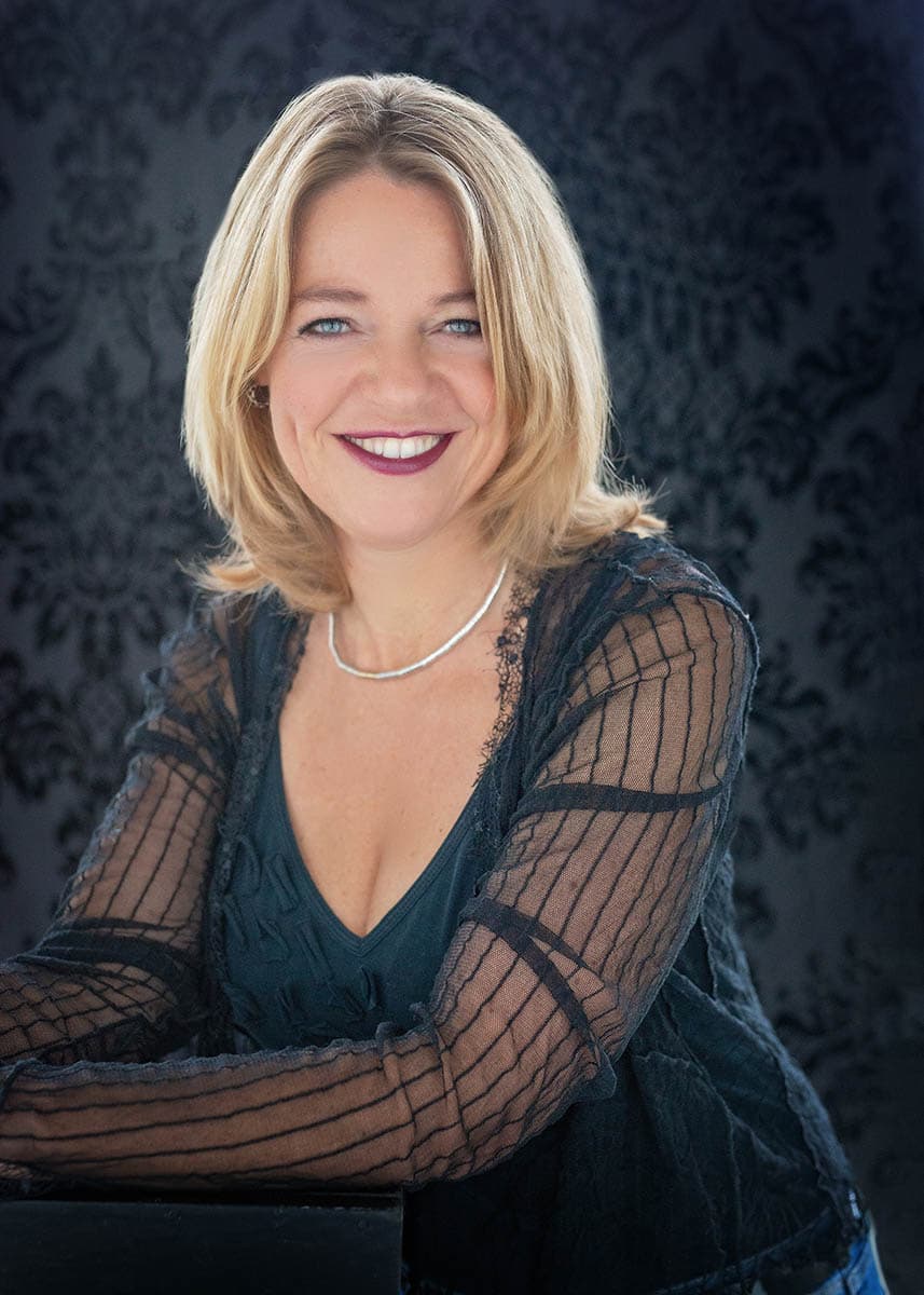 Smiling blonde woman with blue eyes wearing a black sheer striped blouse and a silver necklace posing against a dark patterned background.