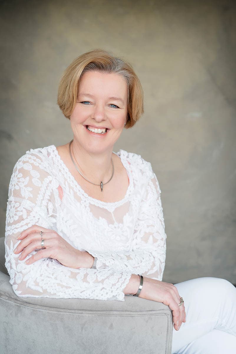 Smiling middle-aged woman with short blonde hair wearing a white lace top and white pants, seated on a gray chair against a neutral background.