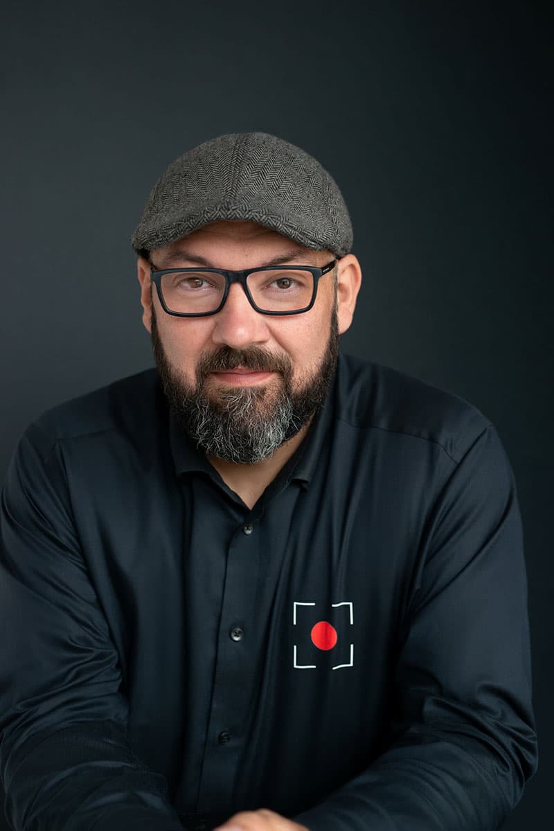 Bearded man wearing glasses, a gray herringbone flat cap, and a black button-up shirt with a red circle and white frame logo, against a dark background.