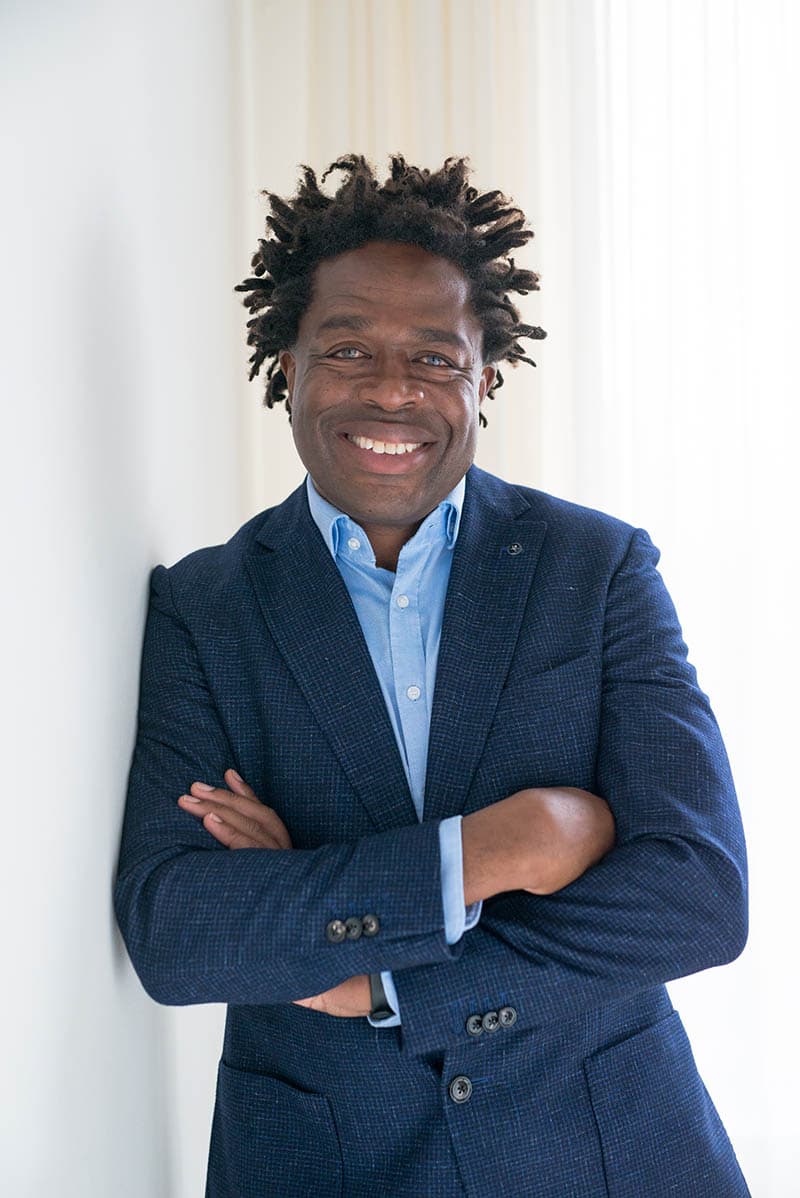 Smiling man with dreadlocks wearing a blue blazer and light blue shirt, standing with arms crossed against a white wall.