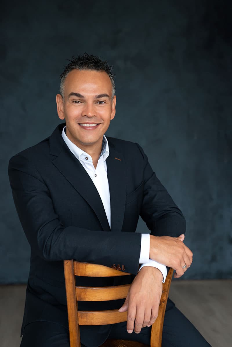 Smiling man in a black suit and white shirt sitting backwards on a wooden chair against a dark textured background.