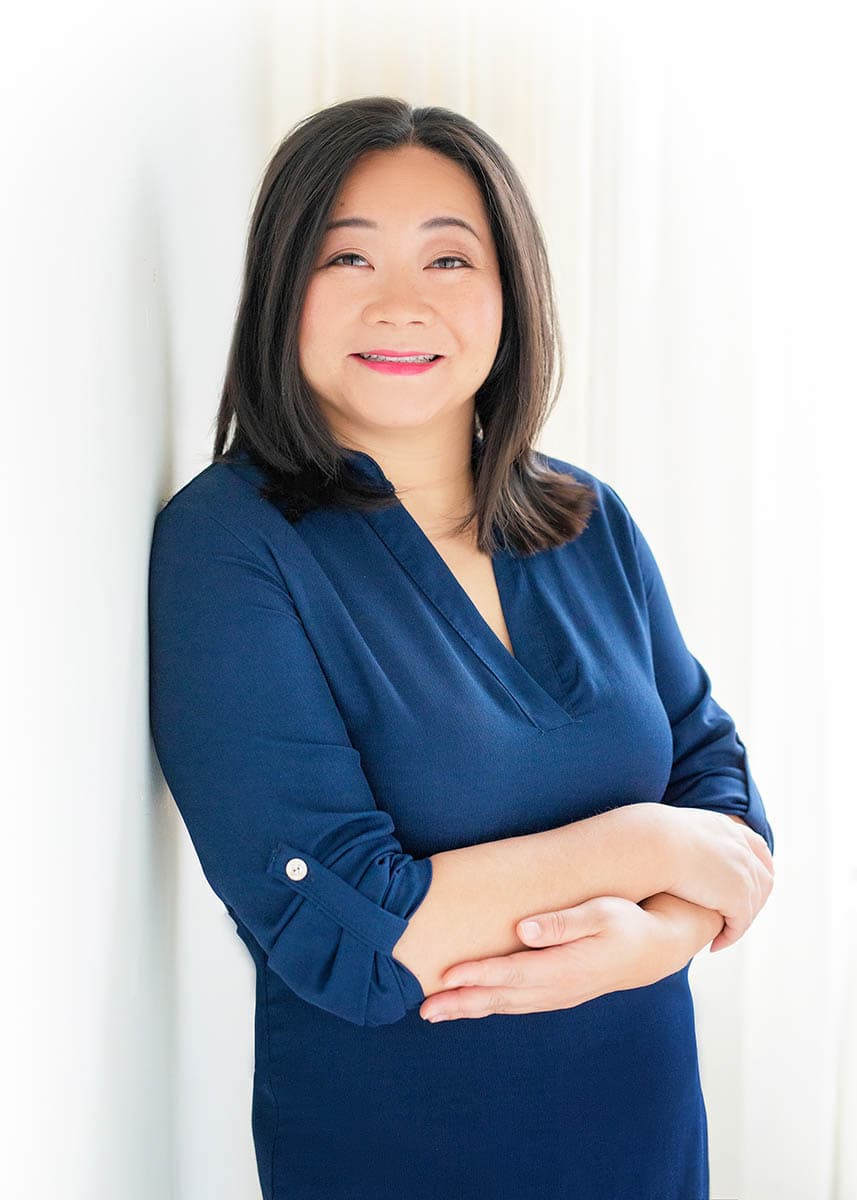 Smiling woman with shoulder-length dark hair wearing a blue long-sleeve top, standing with arms crossed against a white background.