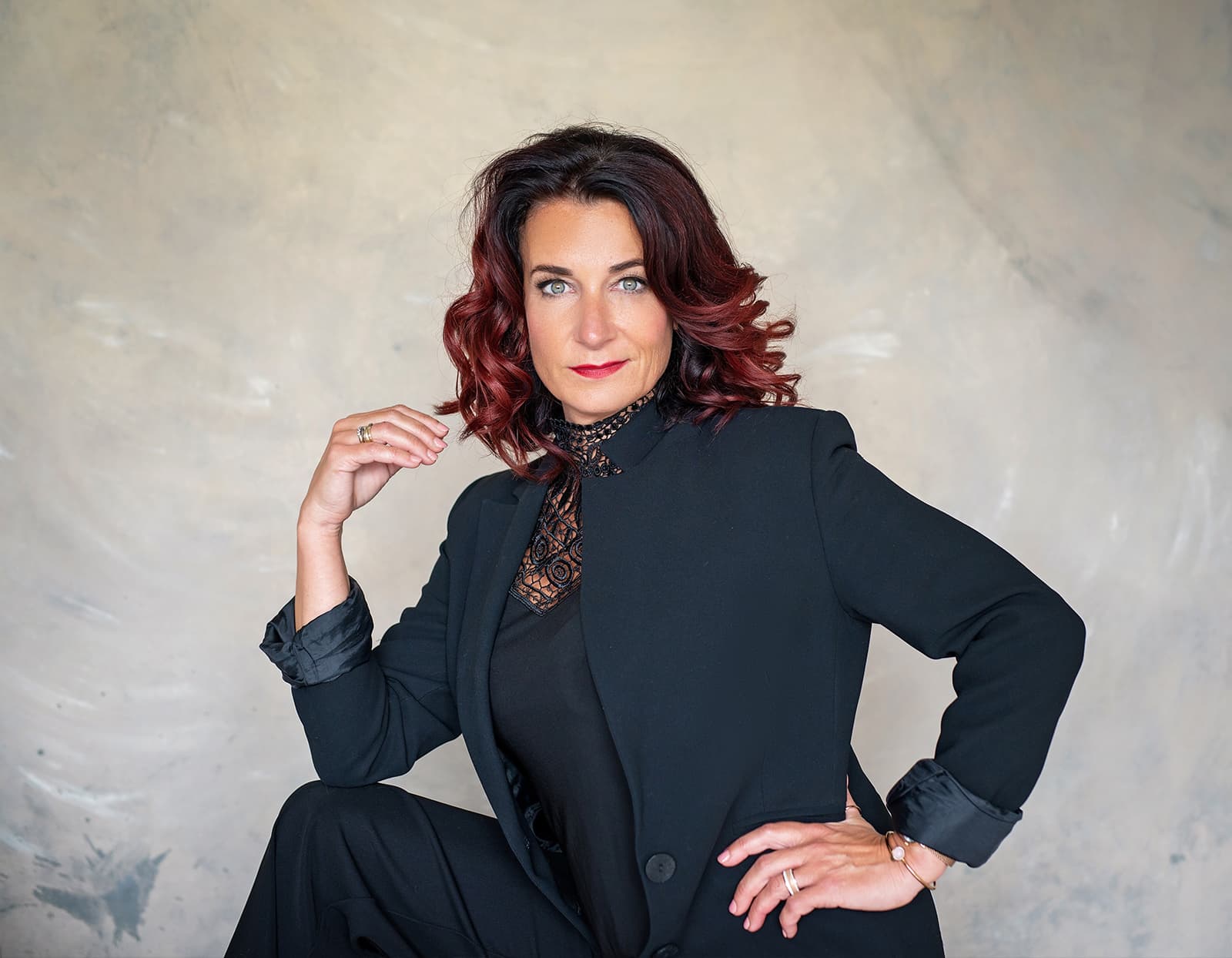 Woman with curly auburn hair wearing a black blazer and lace top poses confidently against a neutral background.