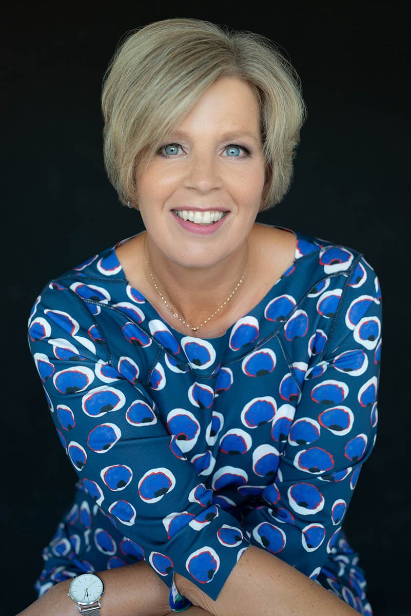 Smiling woman with short blonde hair wearing a blue patterned blouse, a delicate gold necklace, and a silver watch, posed against a dark background.