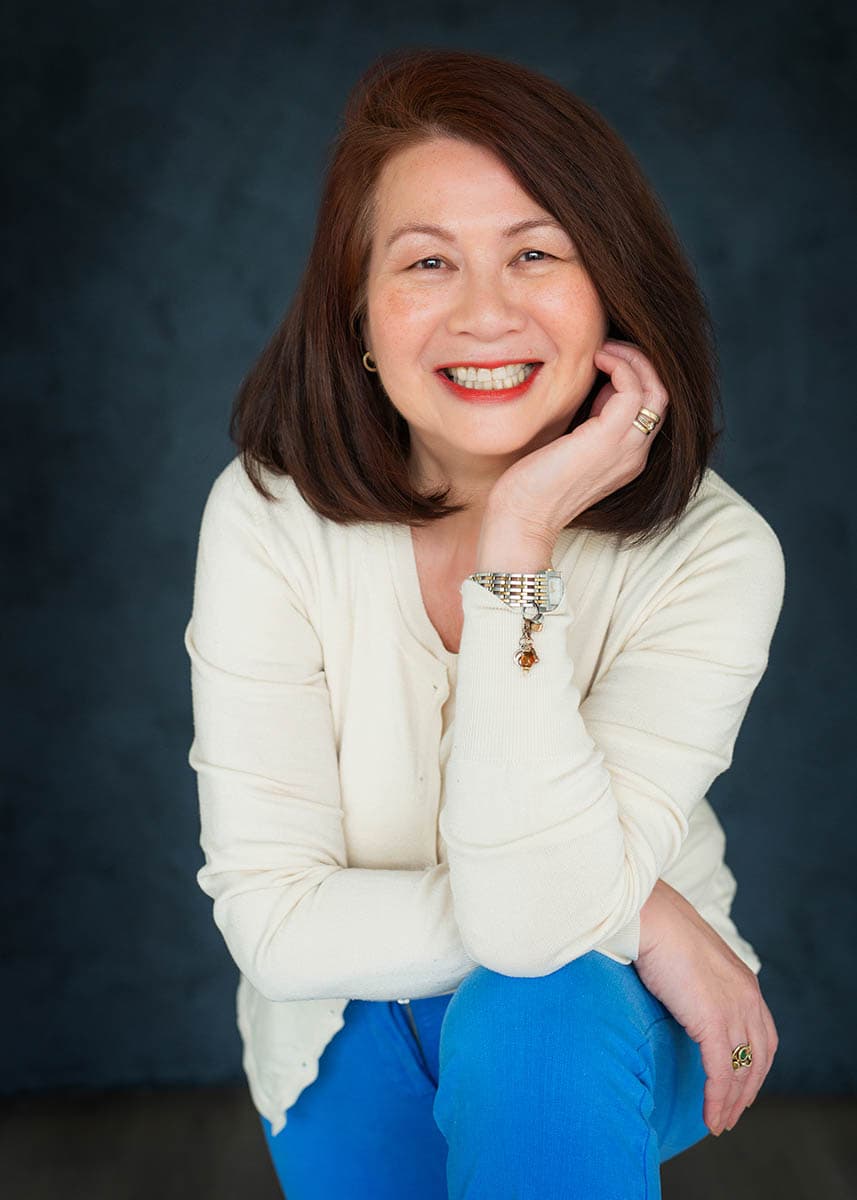 Smiling middle-aged woman with medium-length brown hair, wearing a cream-colored sweater and bright blue pants, resting her chin on her hand against a dark blue background.