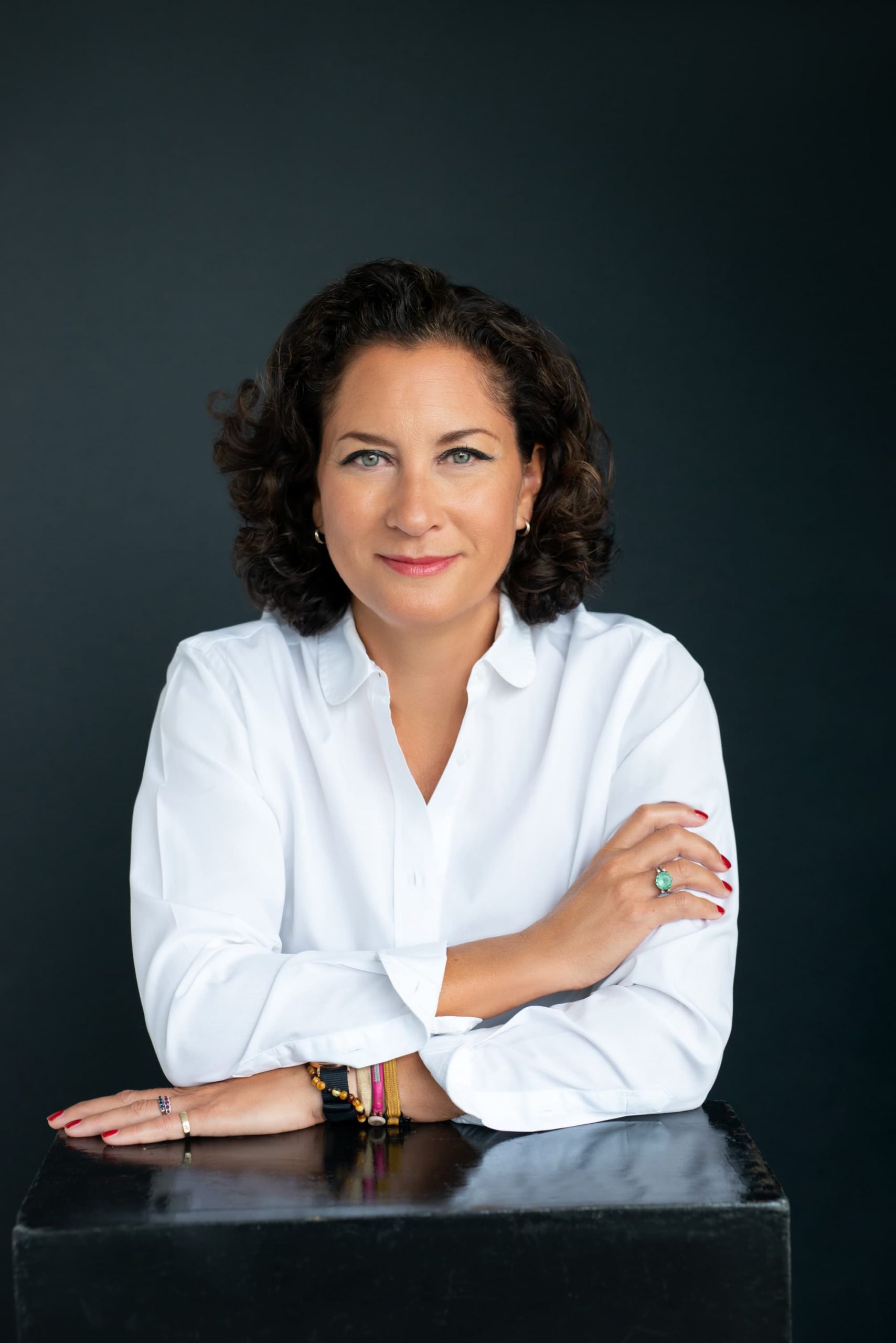 A woman with curly dark hair wearing a white button-up shirt poses against a dark background with a confident expression, resting her folded arms on a black surface.