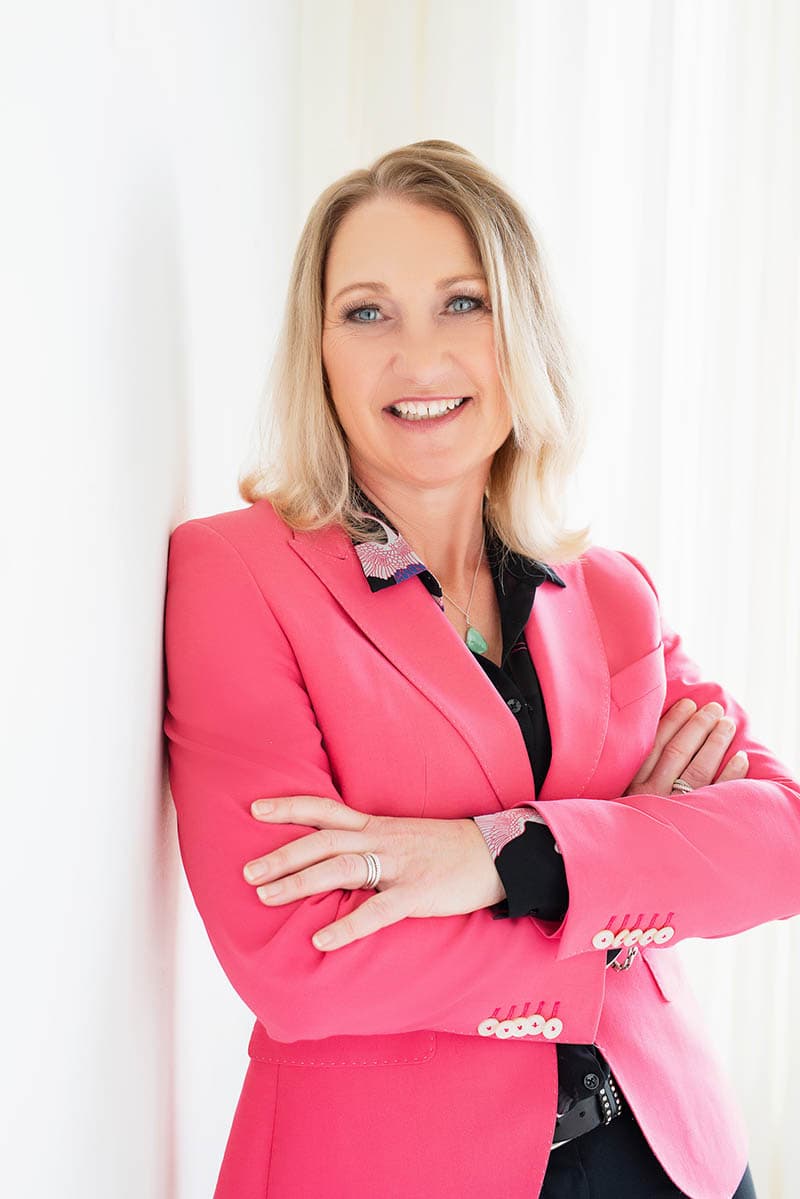 Smiling woman with blonde hair wearing a bright pink blazer over a black shirt, standing with arms crossed against a white wall.