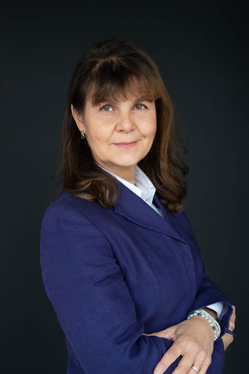 Confident middle-aged woman with brown hair wearing a blue blazer and white shirt, standing against a dark background with arms crossed and a slight smile.