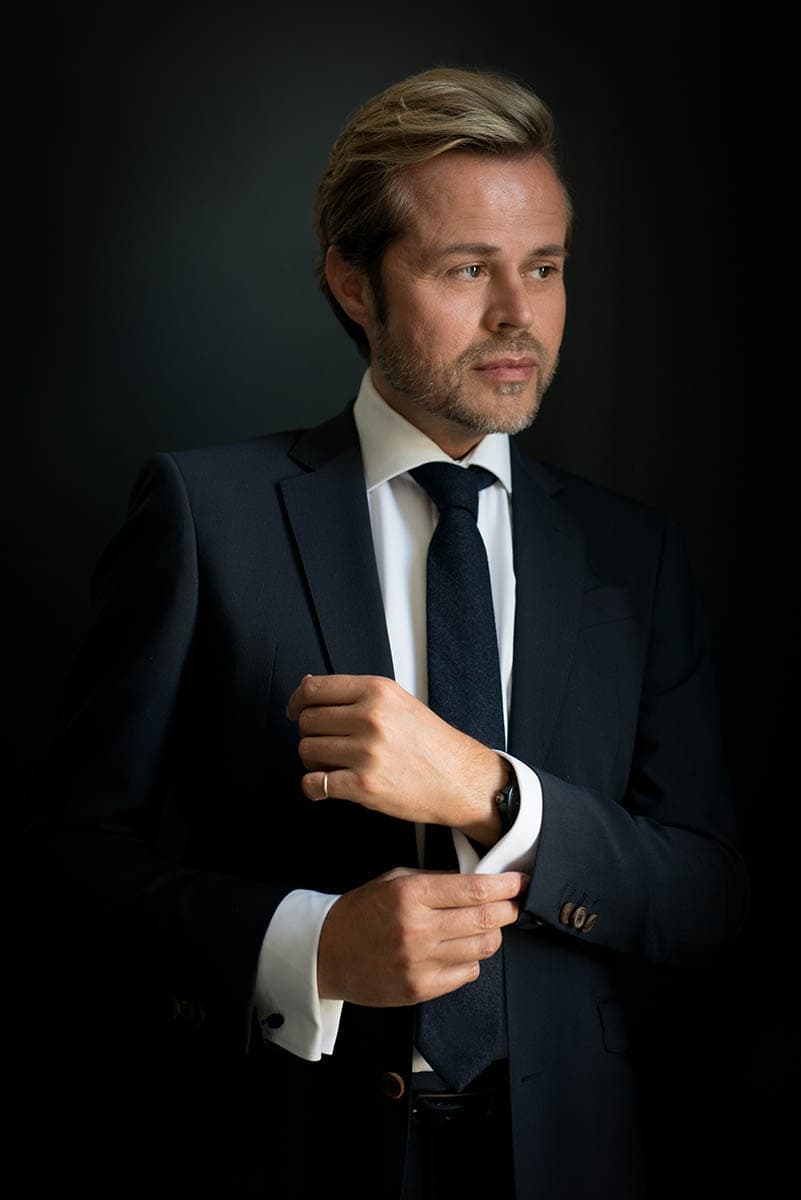 Man with light brown hair and beard wearing a dark suit, white shirt, and dark tie, adjusting his cufflink against a black background.