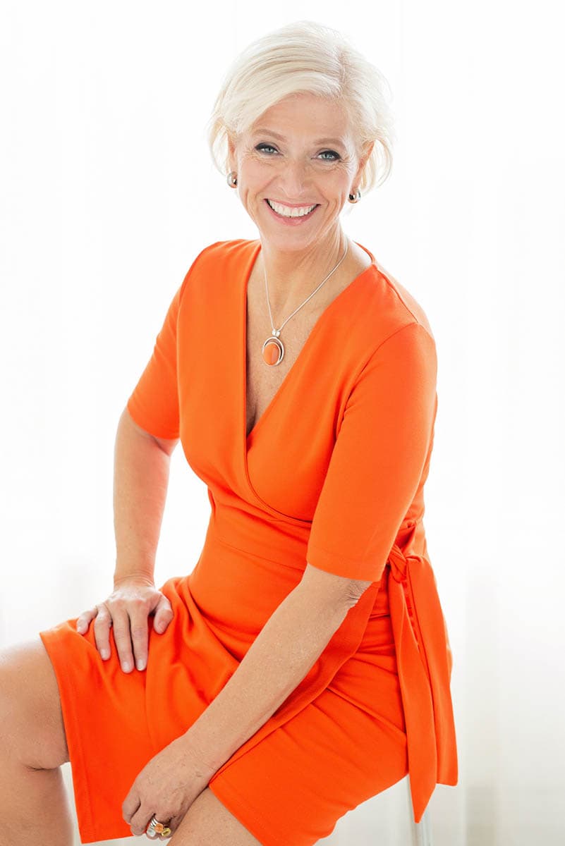 Smiling older woman with short white hair wearing an orange wrap dress, sitting with one hand resting on her knee and the other on her leg, against a bright white background.