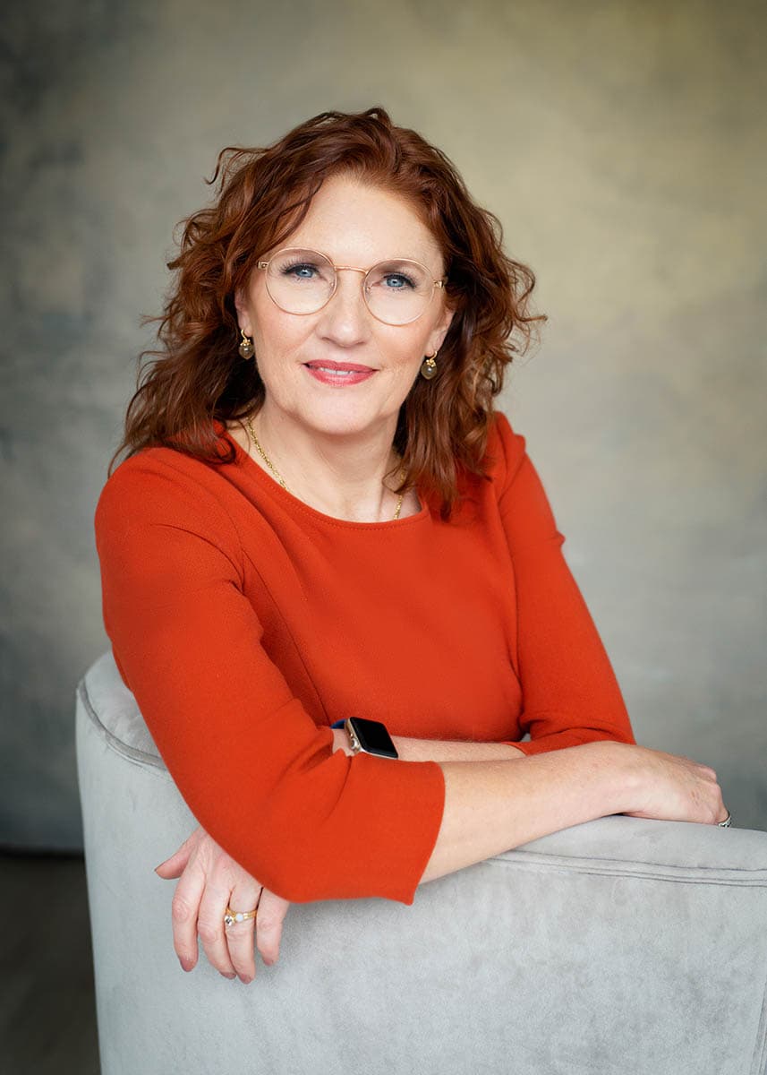 Smiling middle-aged woman with curly red hair and glasses wearing a red top, sitting on a light gray upholstered chair with her arms resting on the back of the chair against a softly blurred gray background.