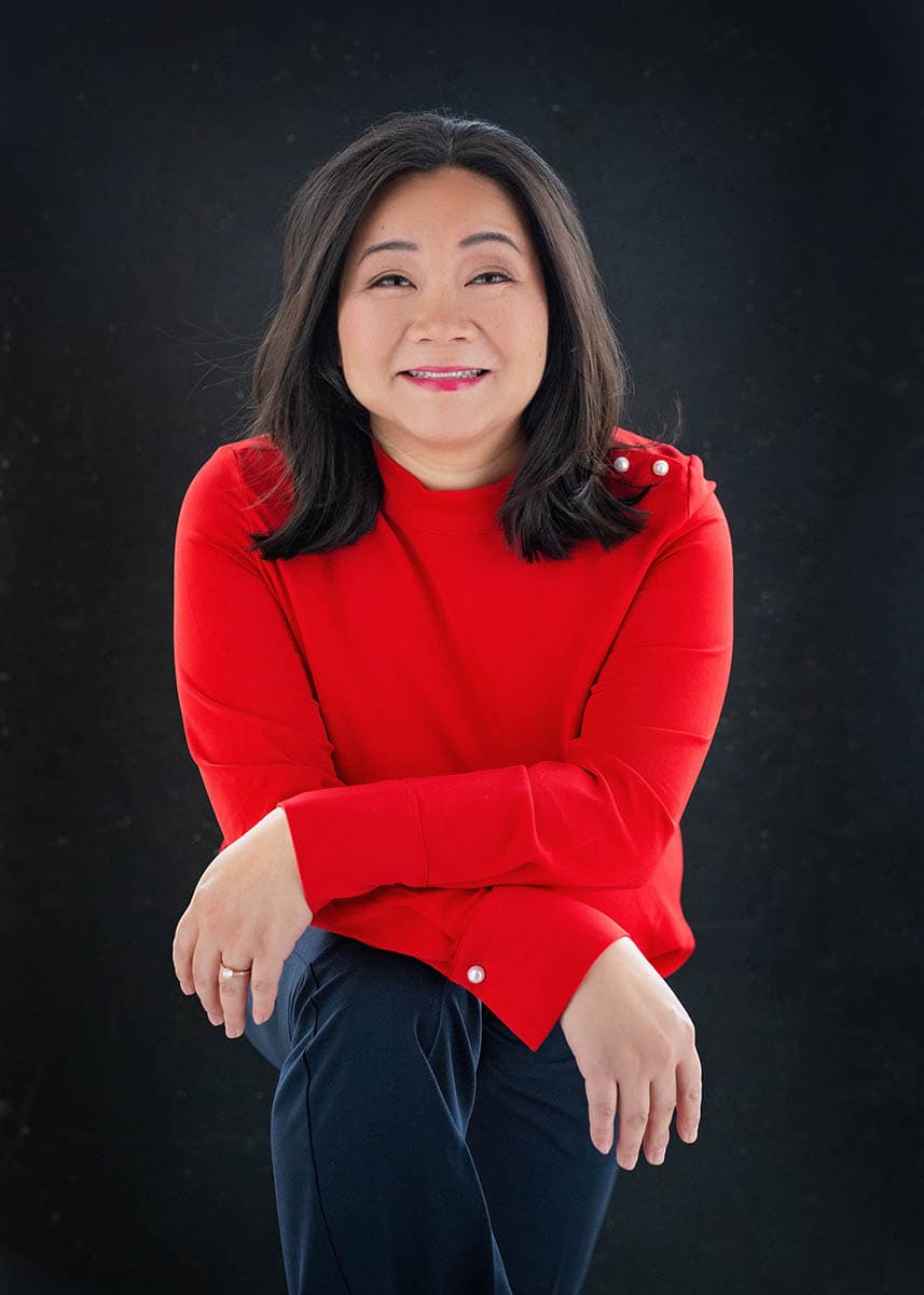 Smiling woman with shoulder-length dark hair wearing a bright red long-sleeve shirt with pearl buttons on the cuffs and black pants, seated against a dark background with arms crossed over one knee.