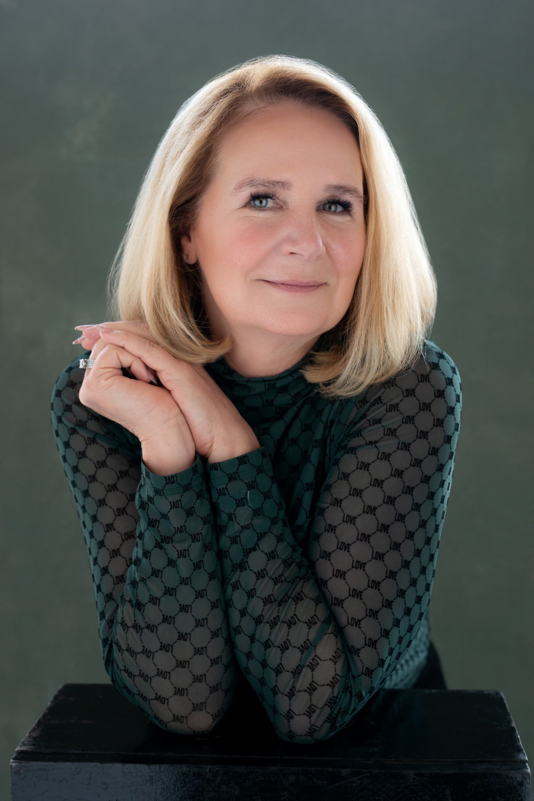 Portrait of a middle-aged woman with blonde hair resting her crossed hands on a black surface, wearing a patterned long-sleeve top with the word 'LOVE' repeated on a dark background.