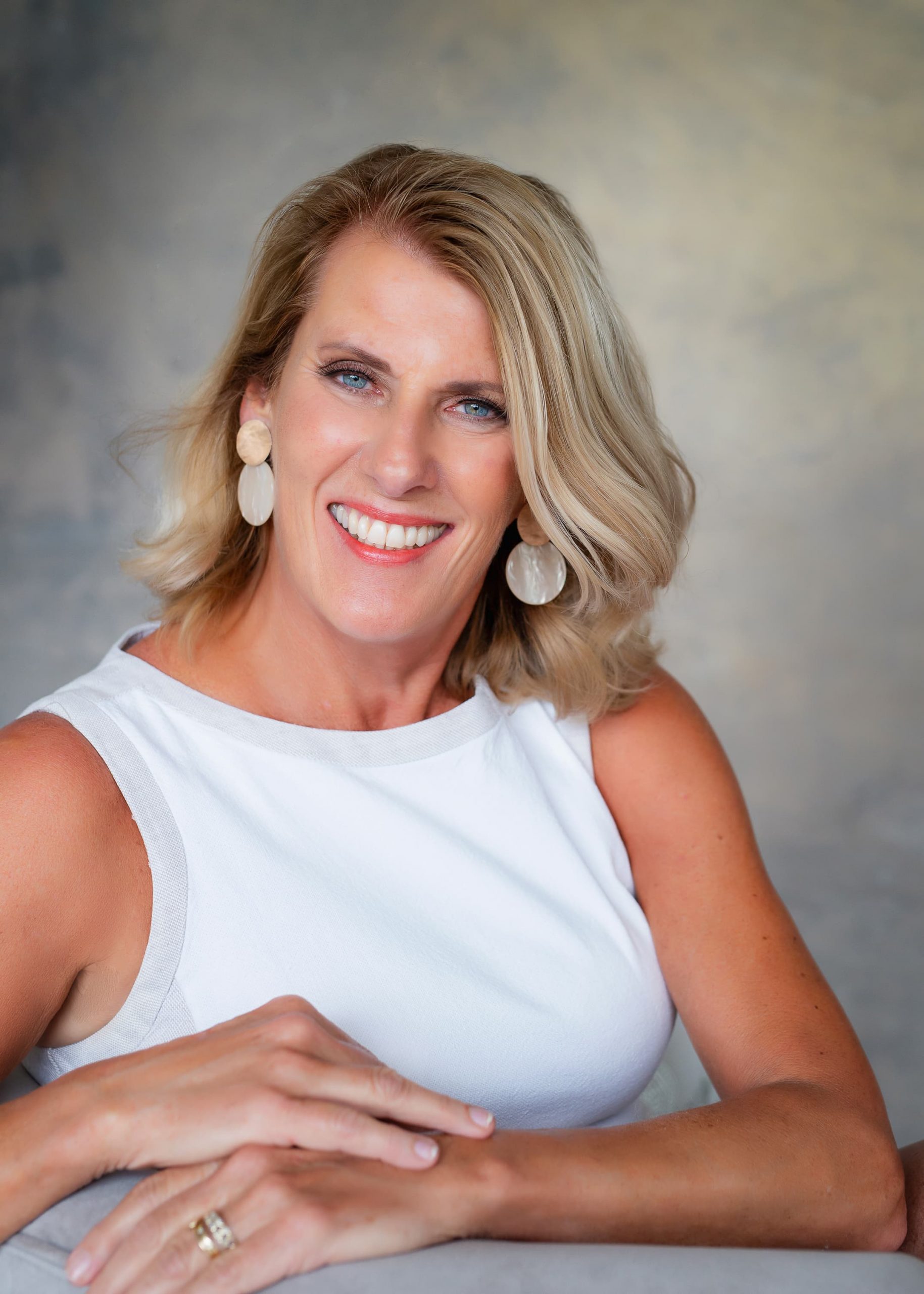 Smiling middle-aged woman with blonde hair and blue eyes wearing a white sleeveless top and large round earrings, resting her hands on a surface.