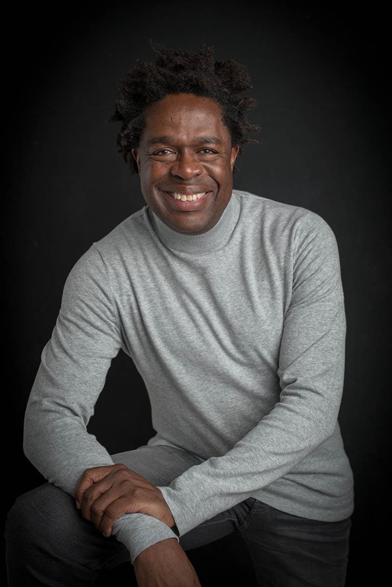 Smiling man with natural curly hair wearing a light gray long-sleeve shirt, posing against a black background.