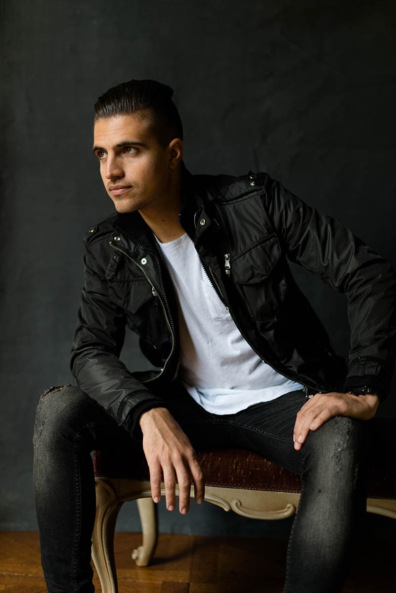 Young man with slicked-back dark hair wearing a black jacket over a white t-shirt and ripped black jeans sitting on a wooden chair against a dark background, looking to the left.