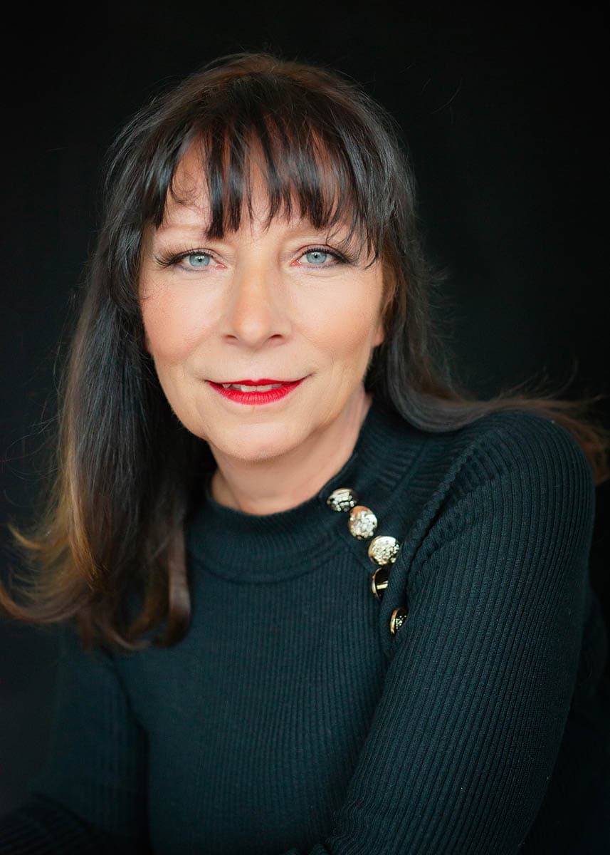 Portrait of a middle-aged woman with light blue eyes and red lipstick wearing a black ribbed sweater with decorative gold buttons on the shoulder against a black background.