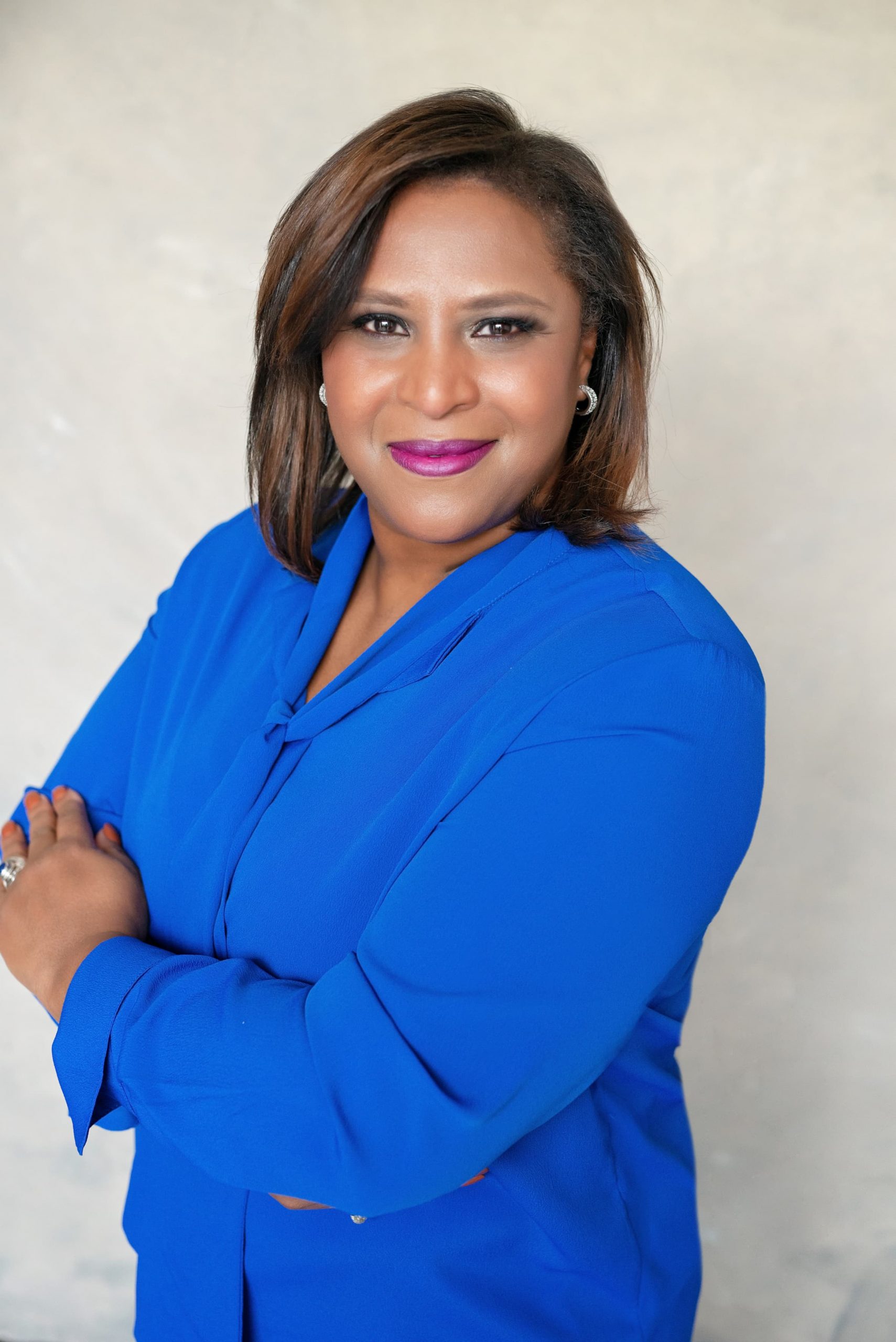 Smiling woman with medium brown hair and purple lipstick wearing a bright blue blouse with arms crossed against a plain light background.