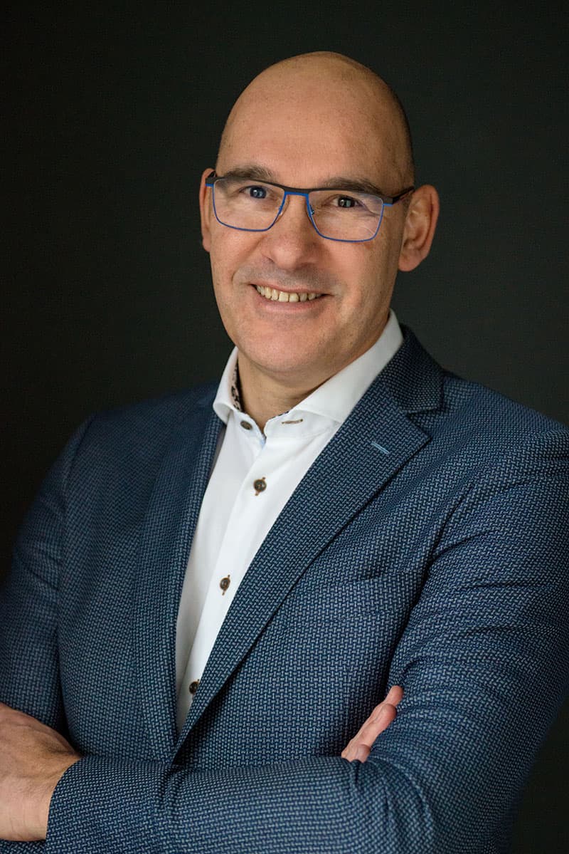 Smiling bald man with glasses wearing a textured navy blue blazer and white shirt, standing with arms crossed against a black background.