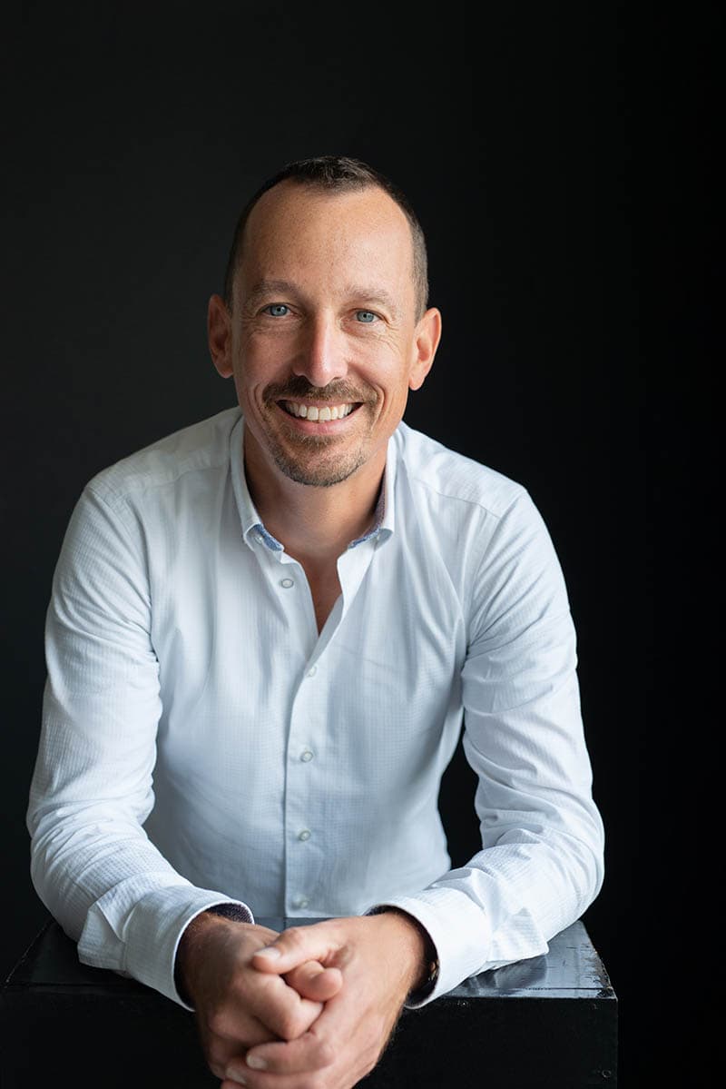 Smiling man with short dark hair and a trimmed goatee wearing a white button-up shirt, sitting and leaning forward with clasped hands against a black background.