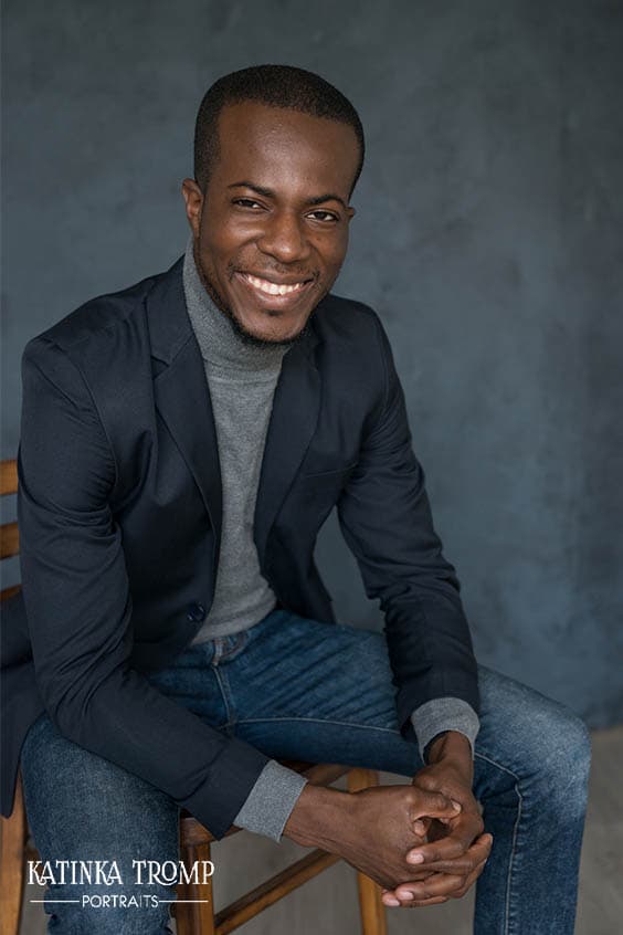 Smiling young man with short hair sitting on a wooden chair against a gray textured background, wearing a dark blazer, gray turtleneck sweater, and blue jeans, hands clasped in front of him.