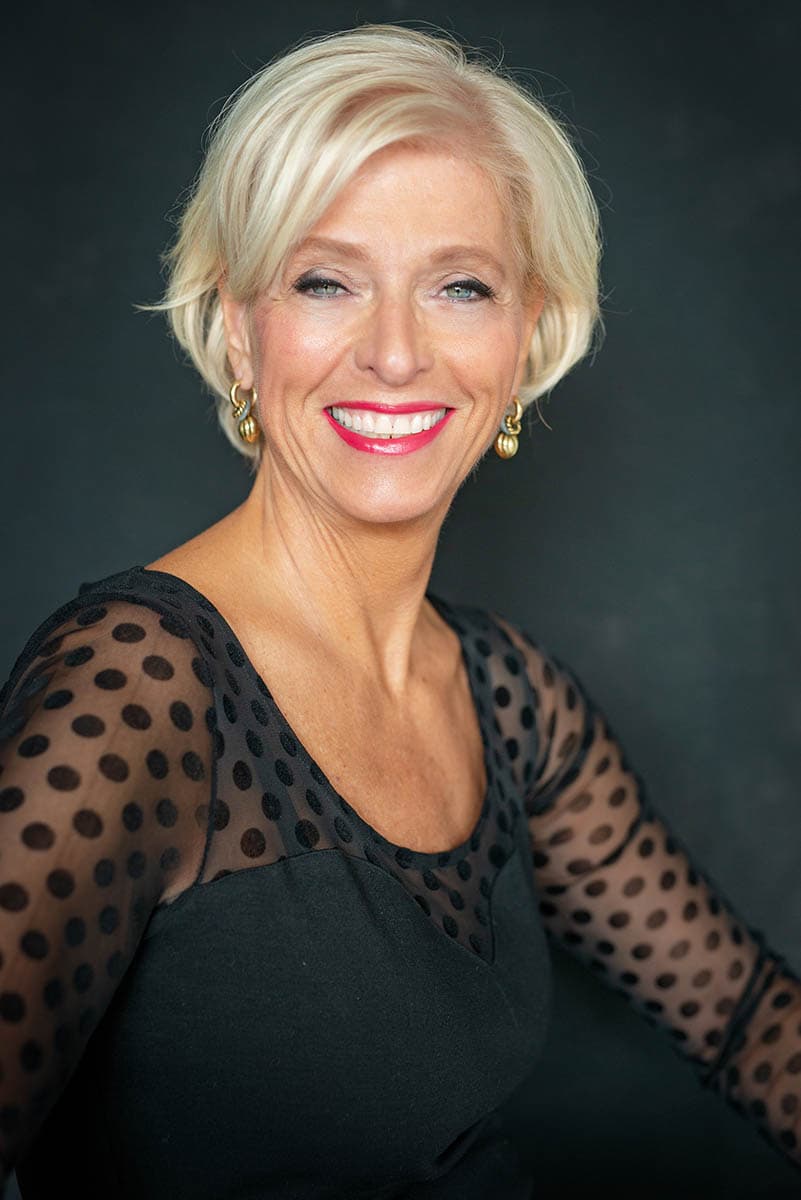 Smiling middle-aged woman with short blonde hair, wearing gold earrings and a black top with sheer polka-dot sleeves against a dark background.