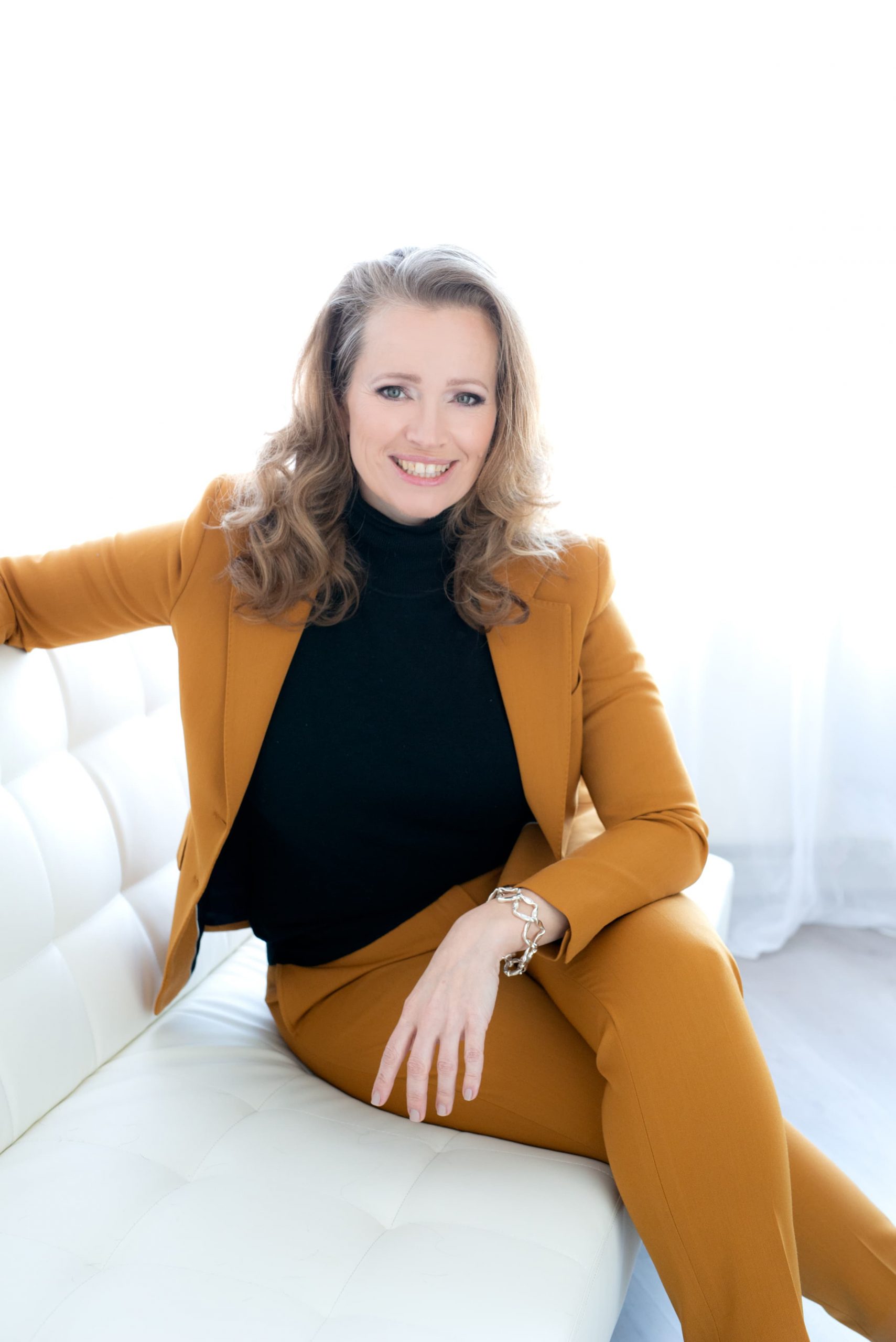 Smiling woman with wavy blonde hair wearing a mustard-colored suit and black turtleneck sitting on a white couch with one arm resting on the back and the other on her knee, wearing a silver bracelet.
