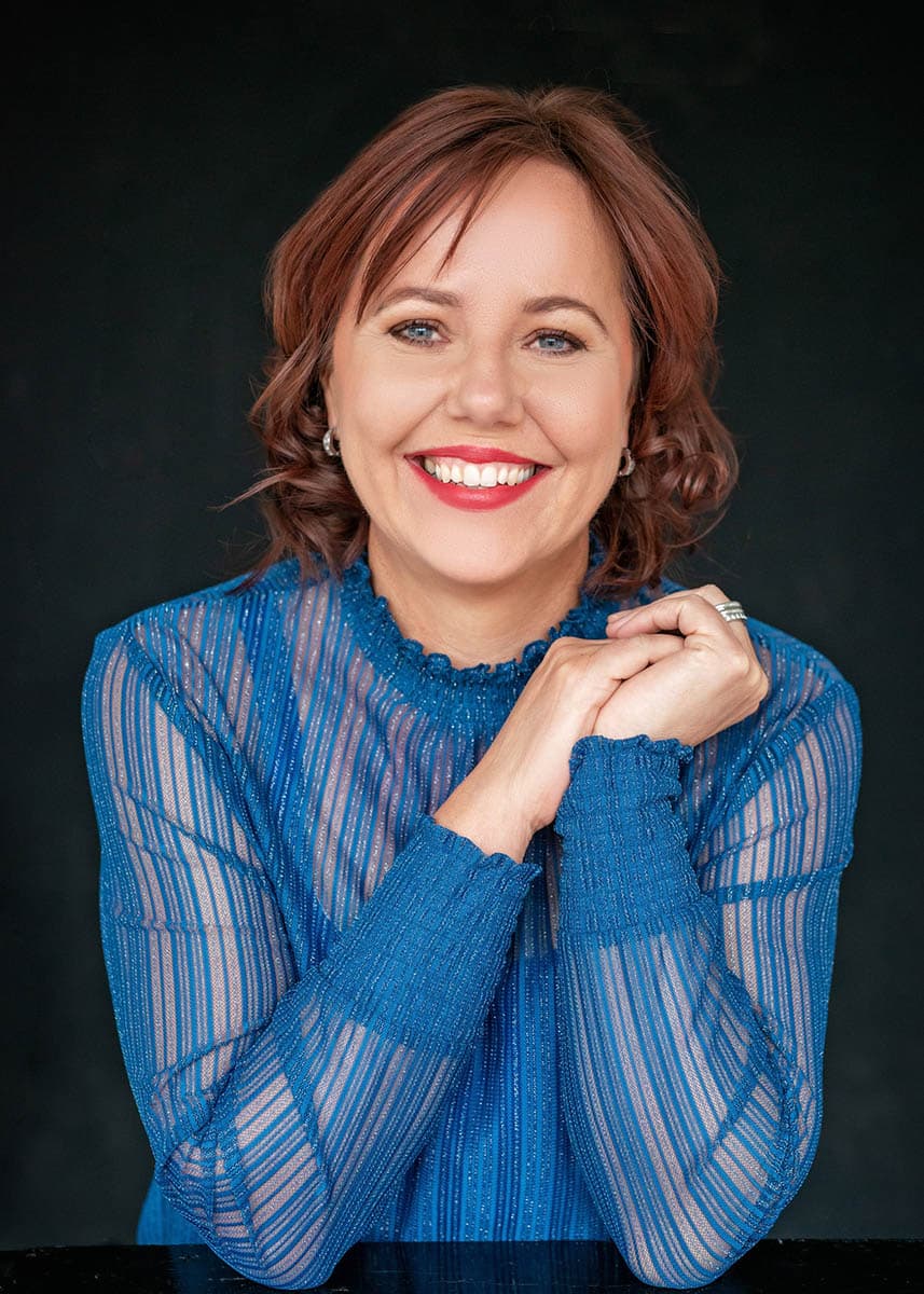 Smiling woman with short auburn hair wearing a sheer blue long-sleeve top, posing with her hands clasped near her chin against a dark background.
