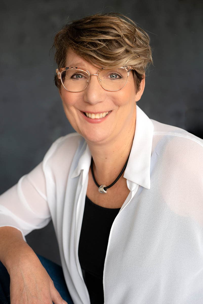 Smiling woman with short wavy hair and glasses wearing a black top, white shirt, and a black necklace with a silver pendant against a dark blurred background.