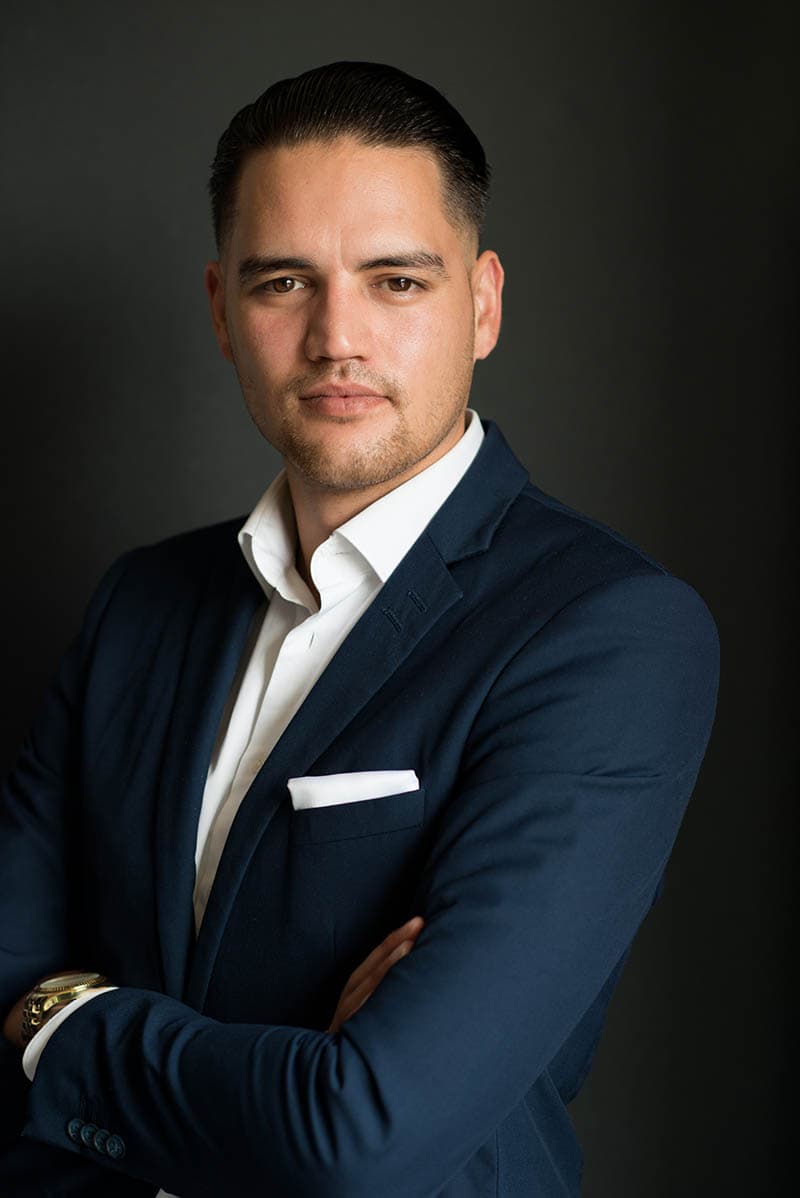 Confident man with slicked black hair wearing a navy suit jacket with a white pocket square and a white dress shirt, standing with arms crossed against a dark background.