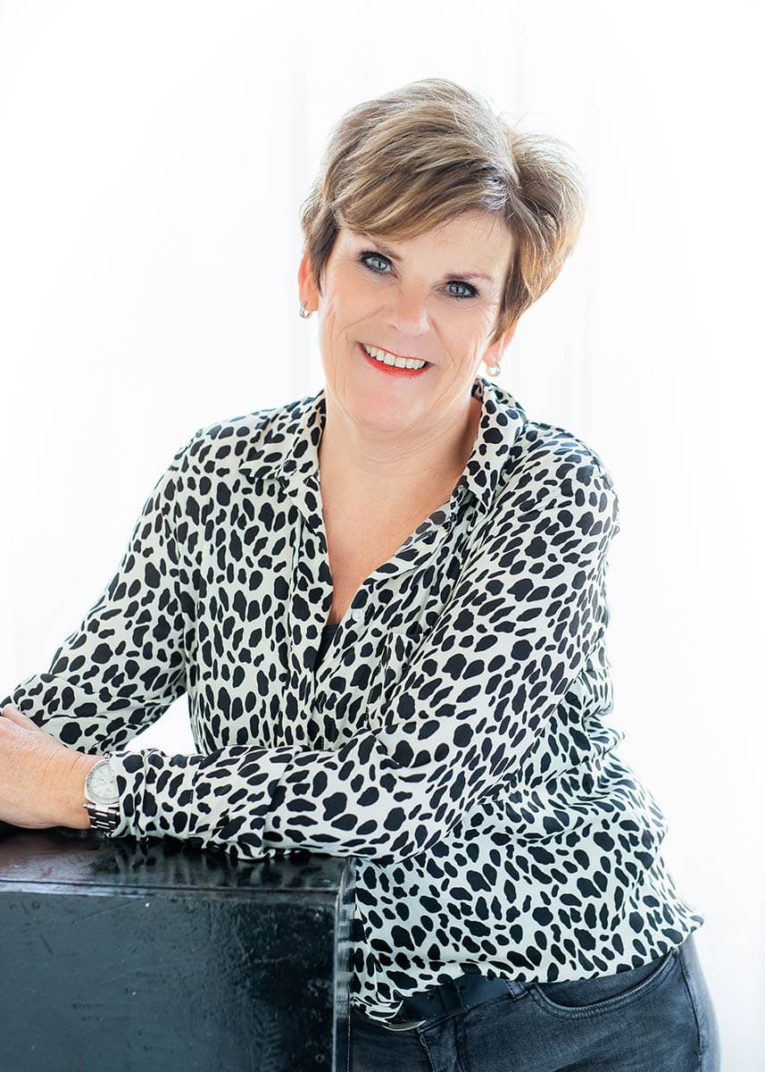 Smiling middle-aged woman with short light brown hair, wearing a white blouse with black spots and a watch, leaning on a black surface.