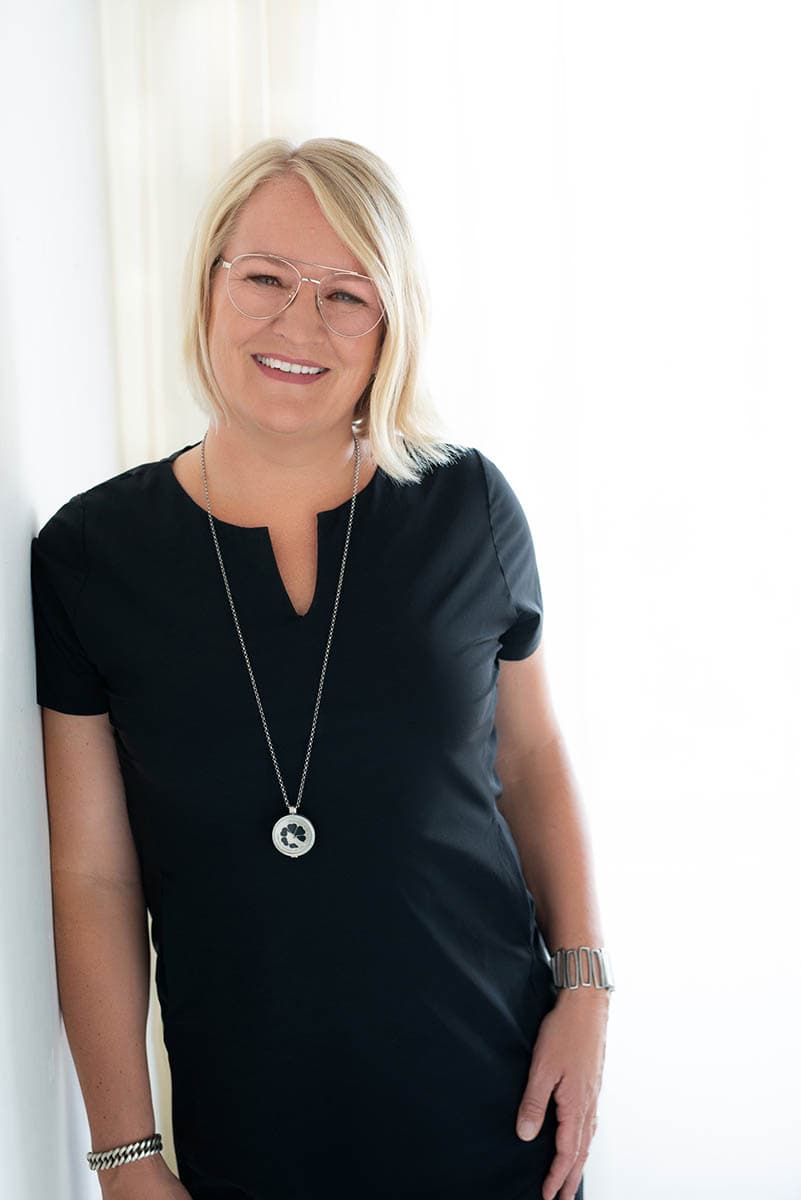 Smiling woman with short blonde hair wearing glasses, a black shirt, a long necklace with a circular pendant, and bracelets, standing against a white background.