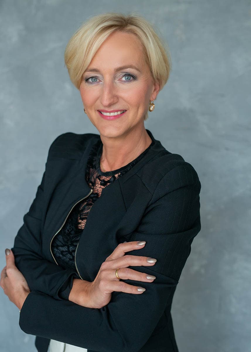 Portrait of a confident middle-aged woman with short blonde hair wearing a black blazer and lace top, smiling with arms crossed against a gray textured background.