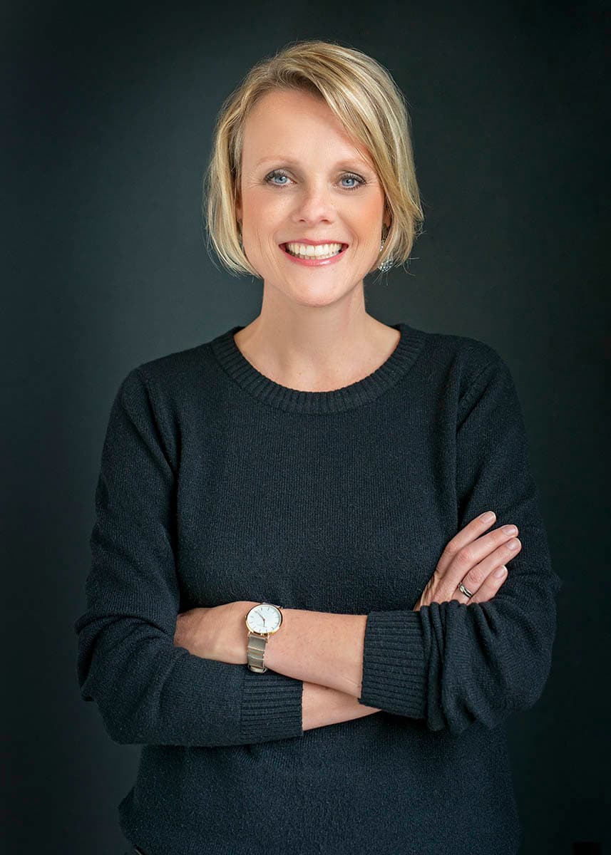 Smiling woman with short blonde hair wearing a black sweater and a watch on her left wrist, standing with arms crossed against a dark background.