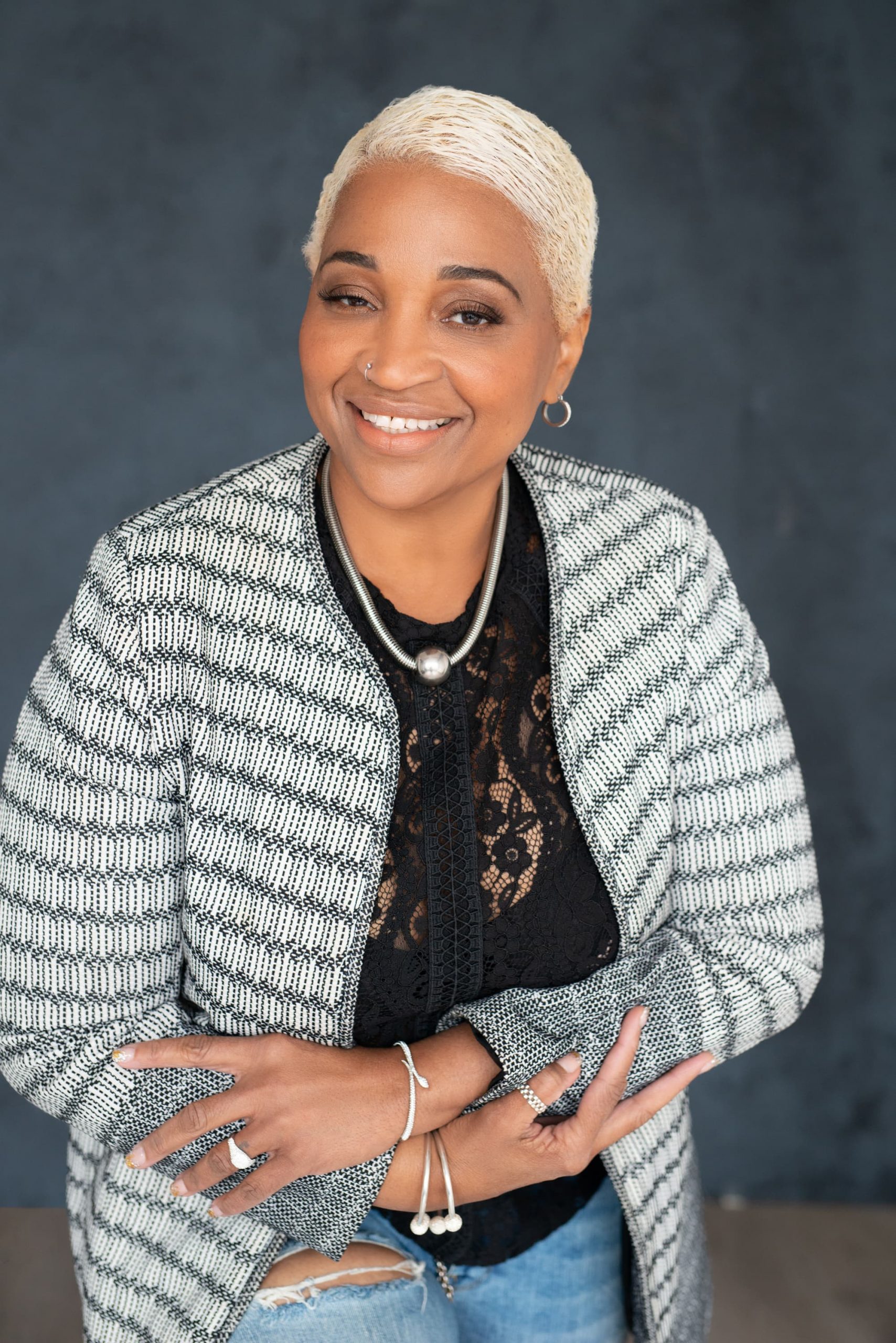 Smiling woman with short platinum blonde hair wearing a black lace top and a patterned gray and white cardigan, accessorized with silver jewelry and light blue ripped jeans, posing against a dark gray background.