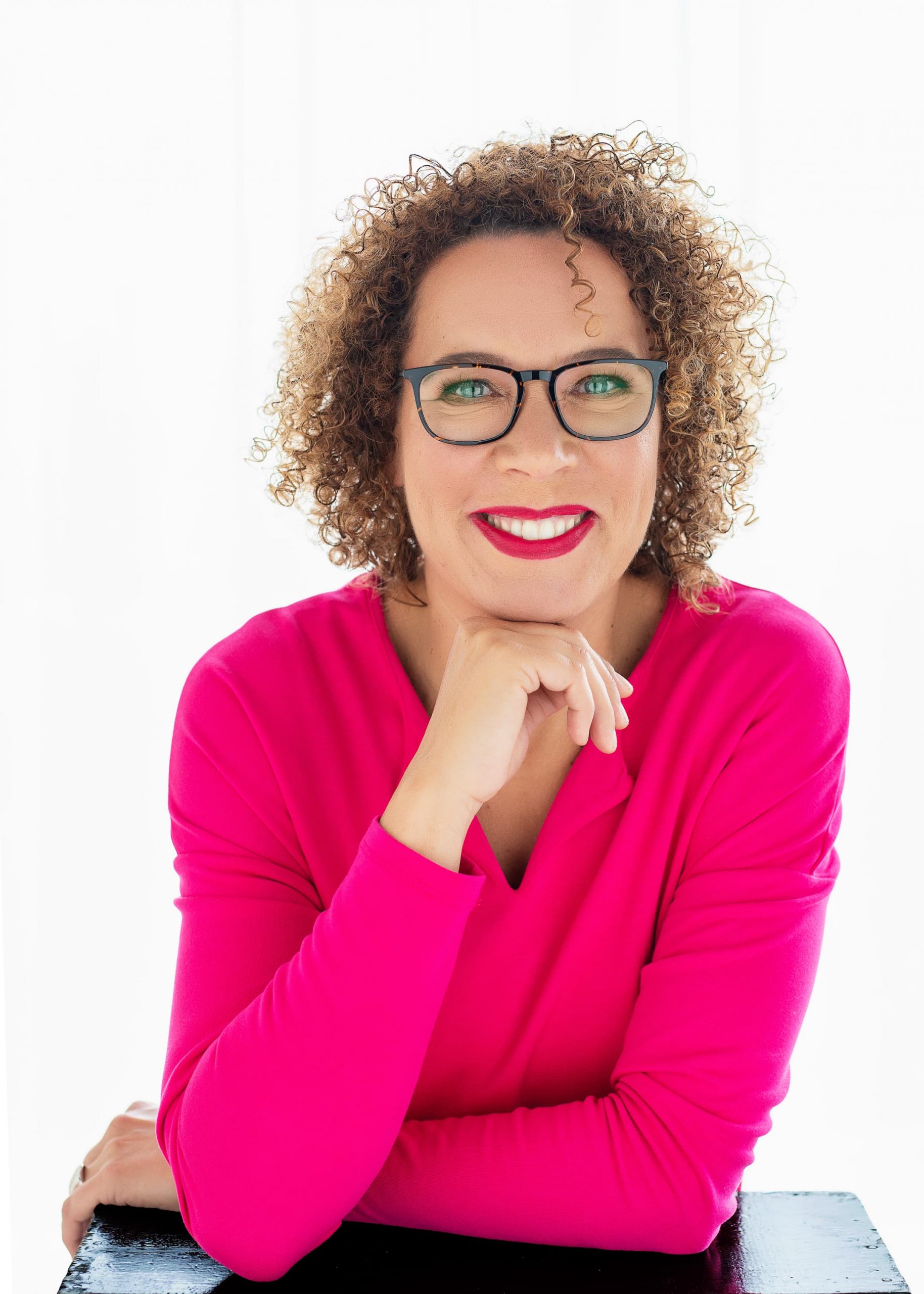 Smiling woman with curly hair and glasses wearing a bright pink long-sleeve top, resting her chin on her hand against a white background.