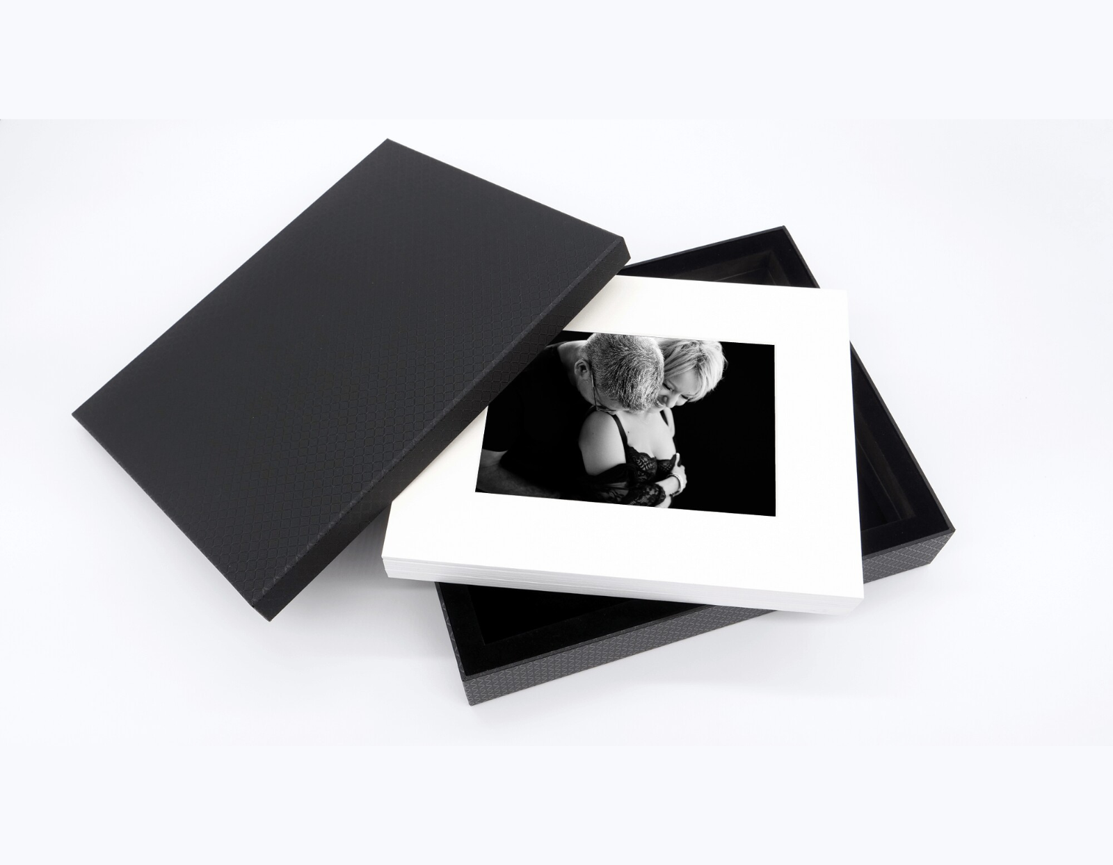 Open black textured box with a stack of white photo prints inside, featuring a black and white intimate portrait of a man embracing a woman dressed in lace.