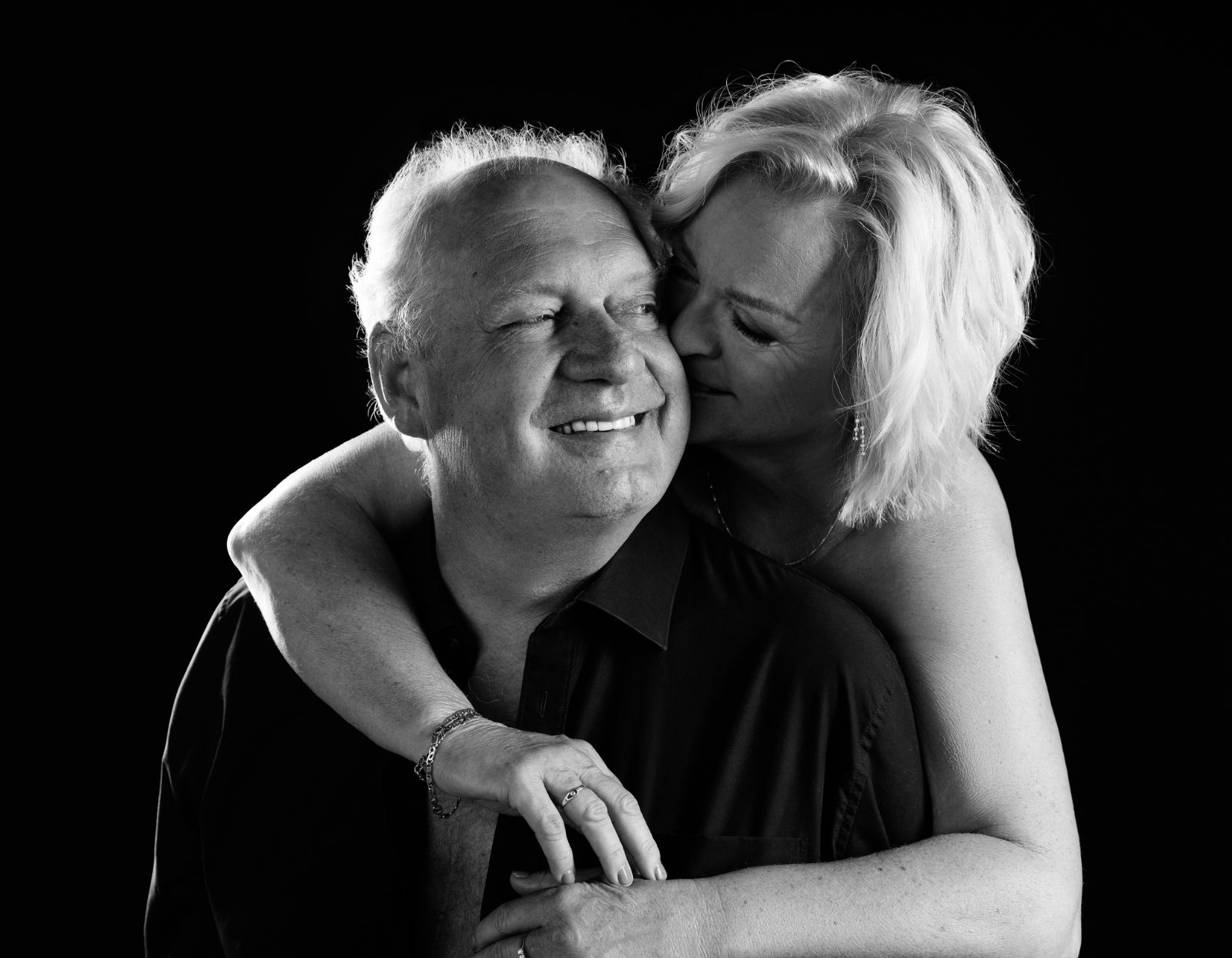Black and white close-up of an older couple embracing, the woman hugging the man from behind and kissing his cheek while he smiles.