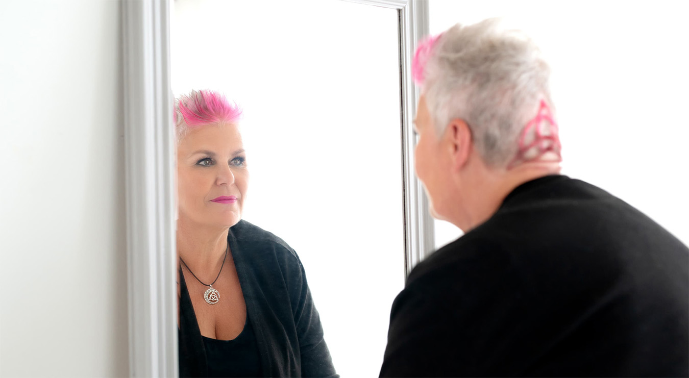 Middle-aged woman with short gray hair styled with bright pink highlights looks at her reflection in a mirror, wearing a black top and necklace with a circular pendant.