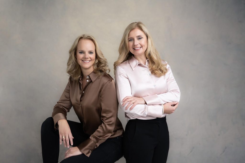 Two smiling women with blonde hair posing against a plain grey background; the woman on the left wears a brown satin blouse and black pants while sitting with one knee up, the woman on the right wears a light pink satin blouse and black pants with her arms crossed.