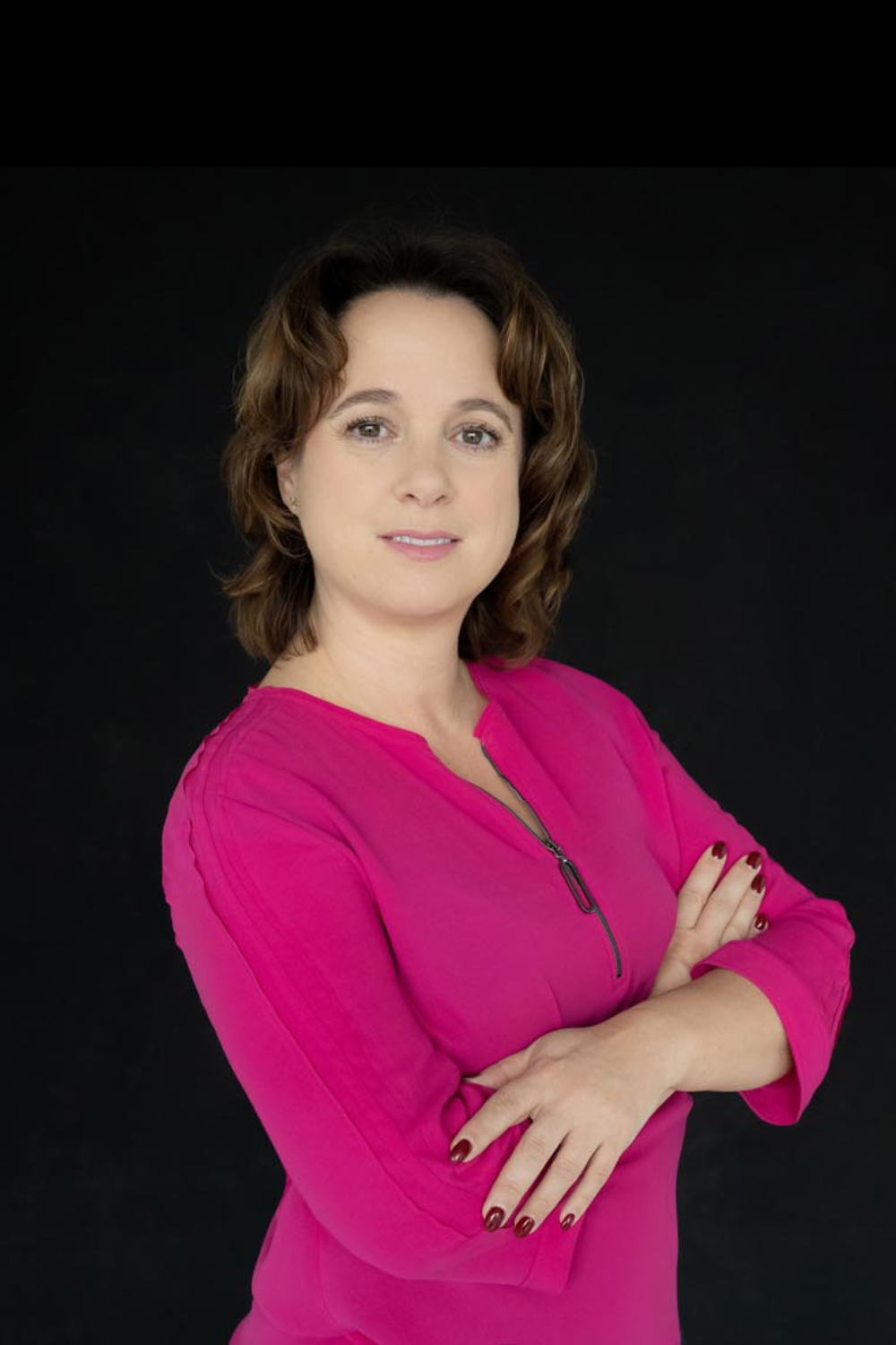 Confident woman with light skin and shoulder-length brown hair, wearing a bright pink blouse with a zipper, arms crossed against a black background.